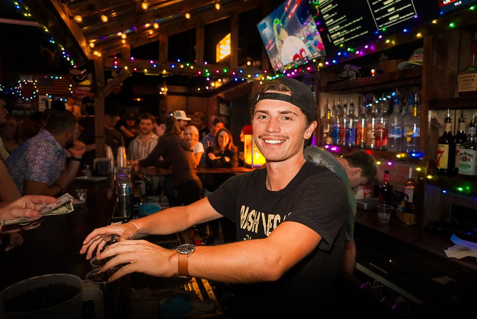Smiling man in a cap behind a bar, lit by colorful string lights in a lively nightclub