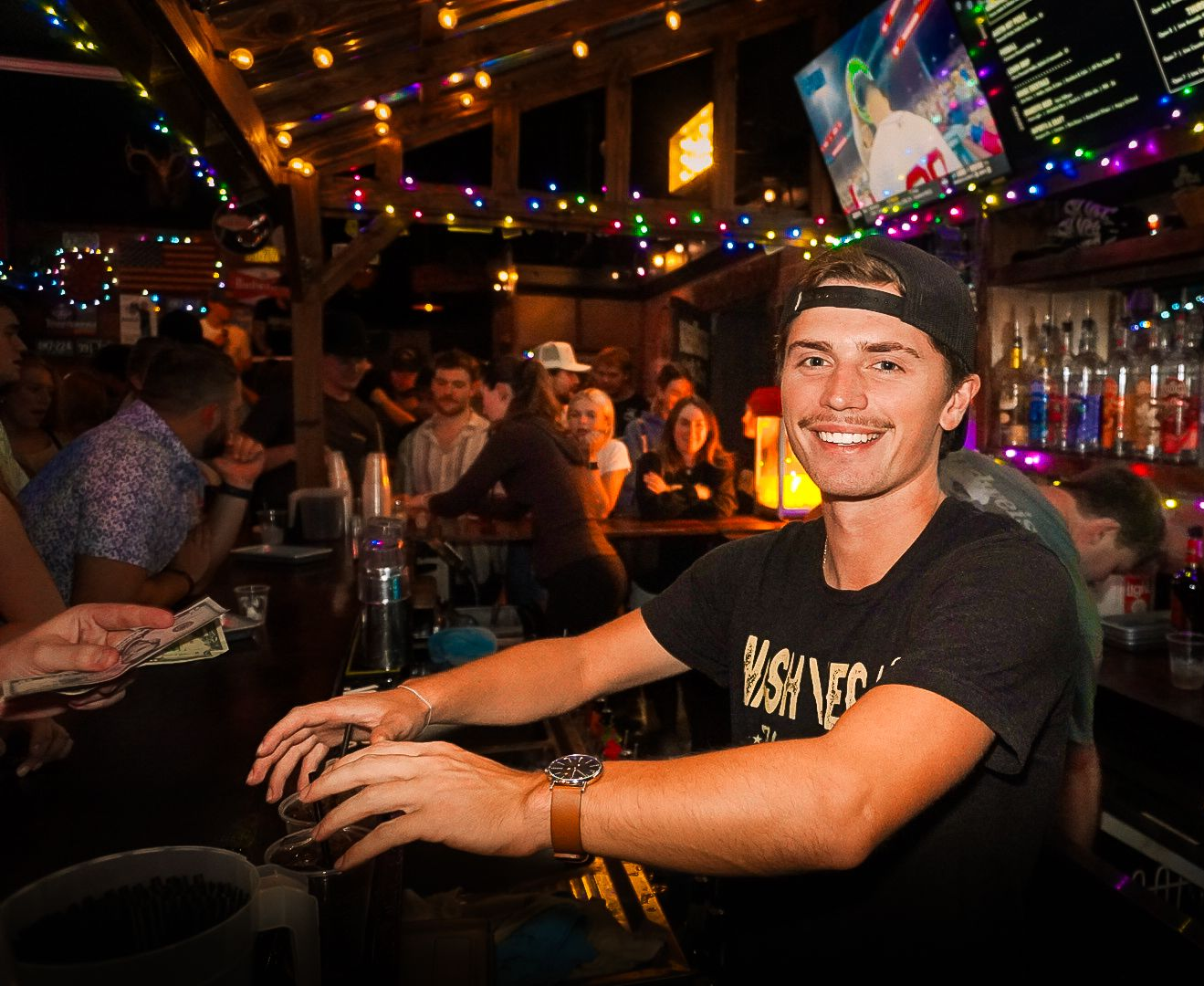 Smiling man in a black T-shirt and cap leaning on a bar in a lively, dimly lit nightclub