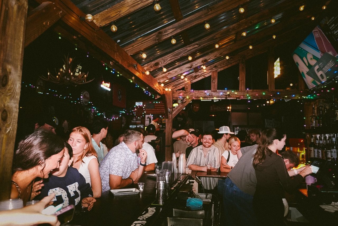 Crowded bar with people chatting under string lights and wooden beams, drinks on tables.