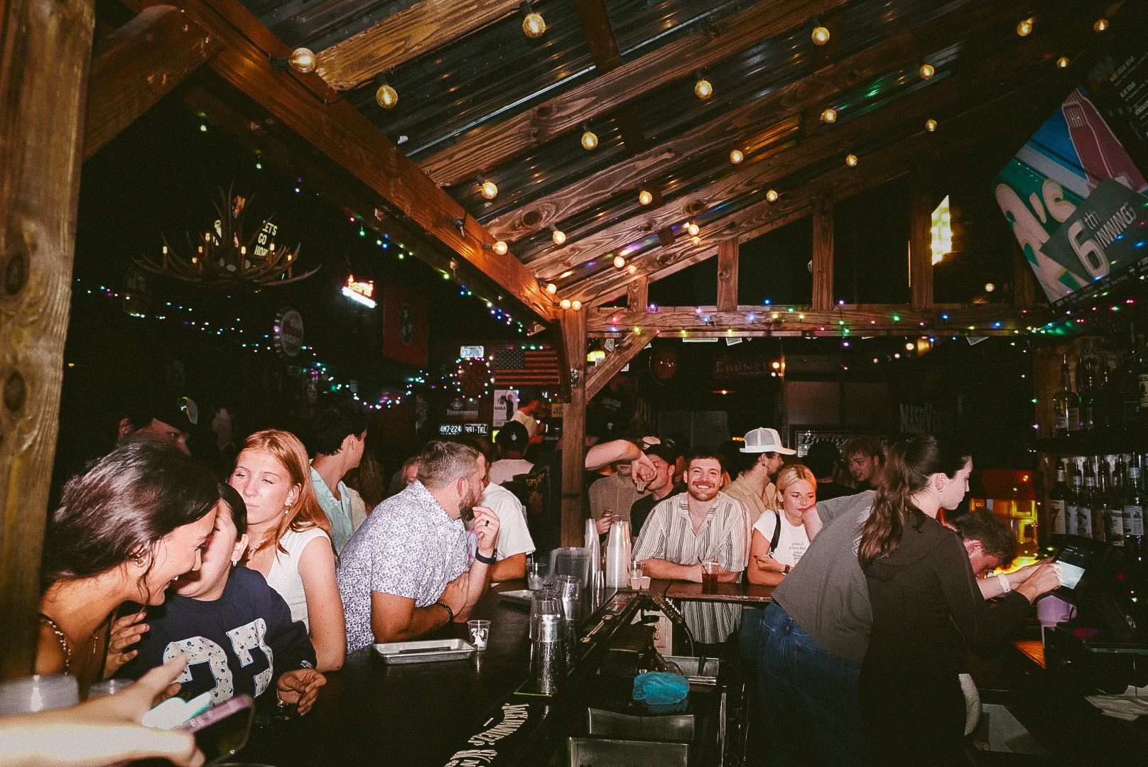 Crowded bar with people chatting under warm string lights and wooden beams