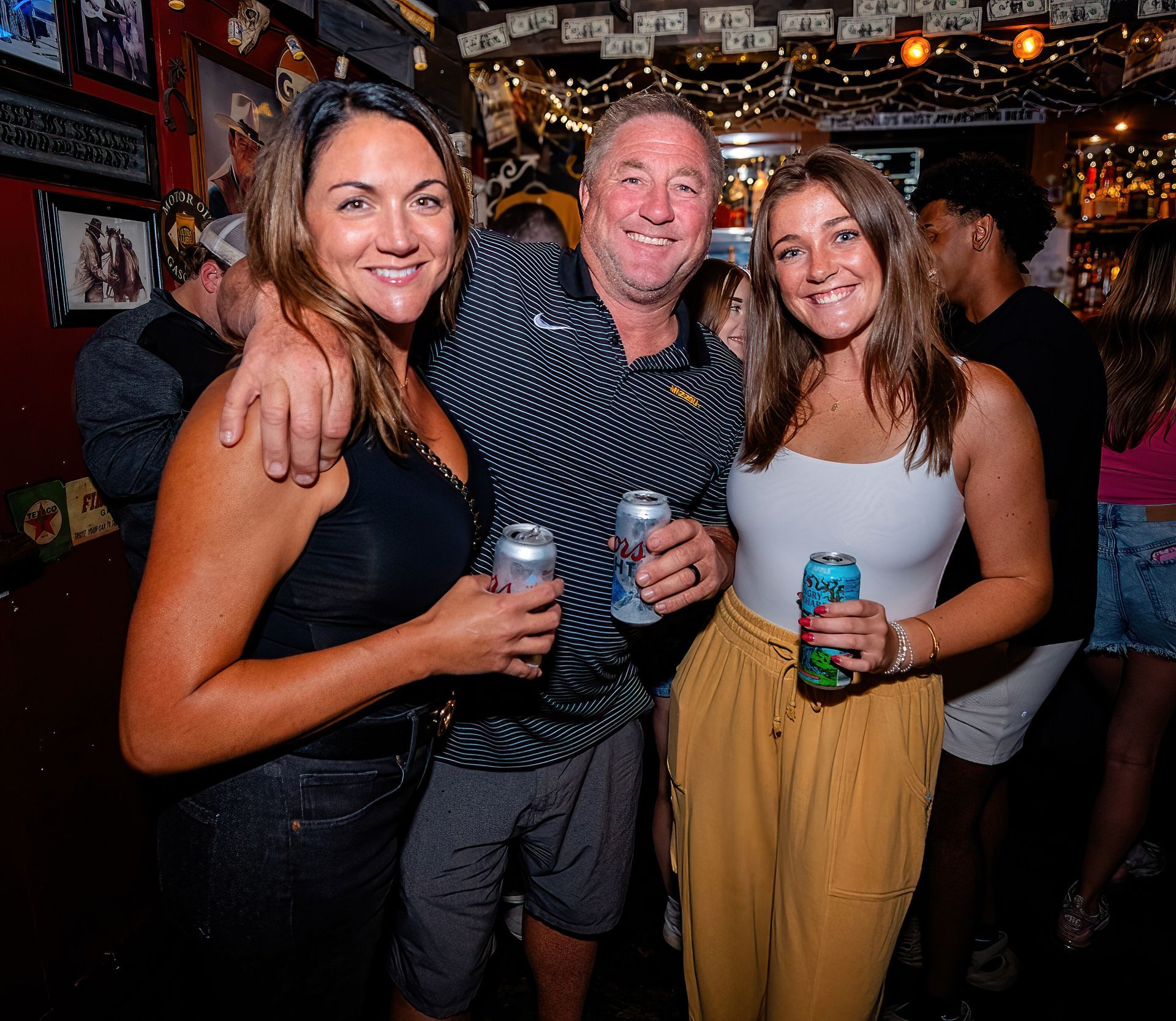 Three people smiling and holding drinks at a lively bar or party scene