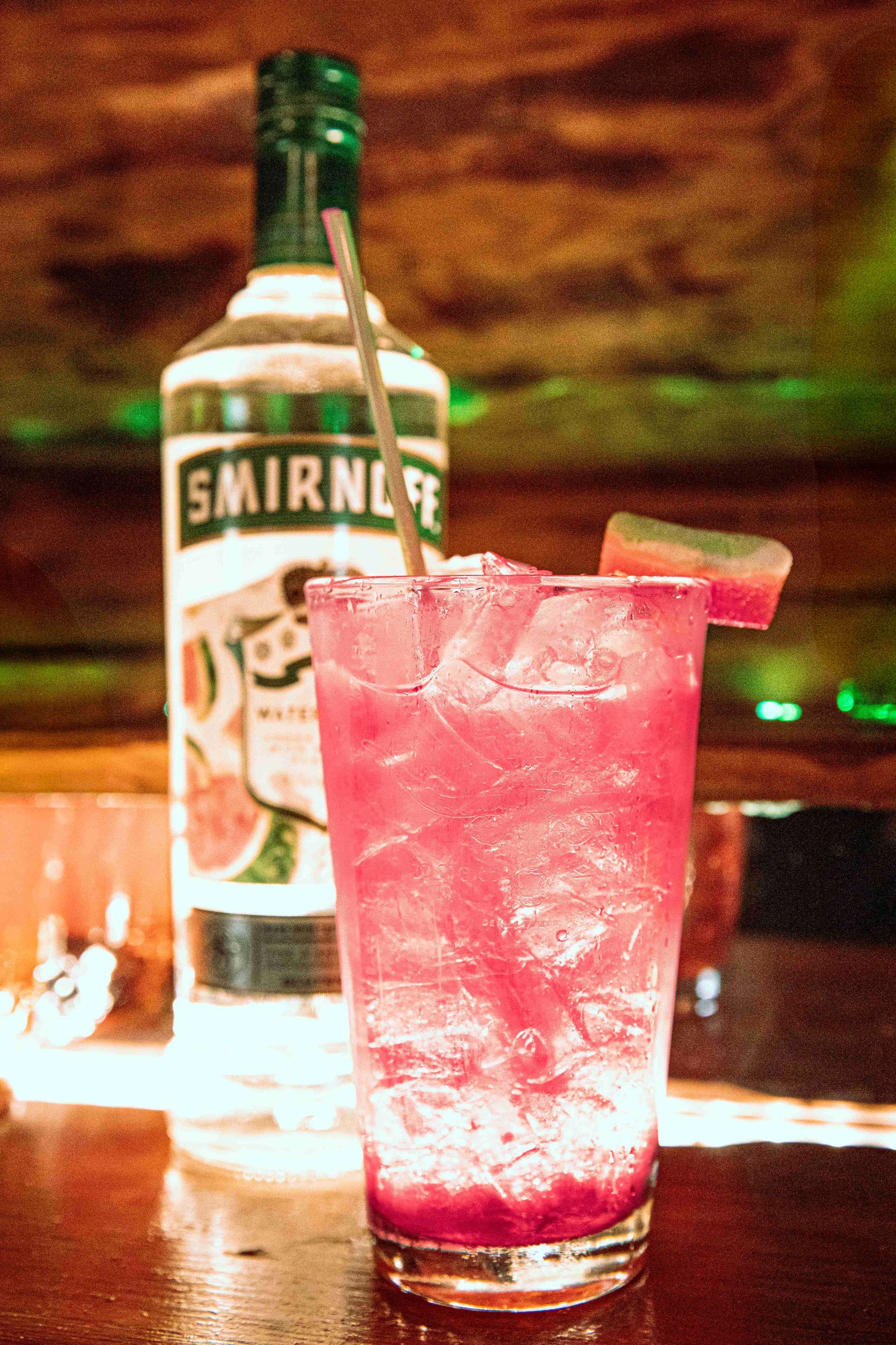 Pink cocktail with ice and lime wedge beside a Smirnoff bottle on a bar counter