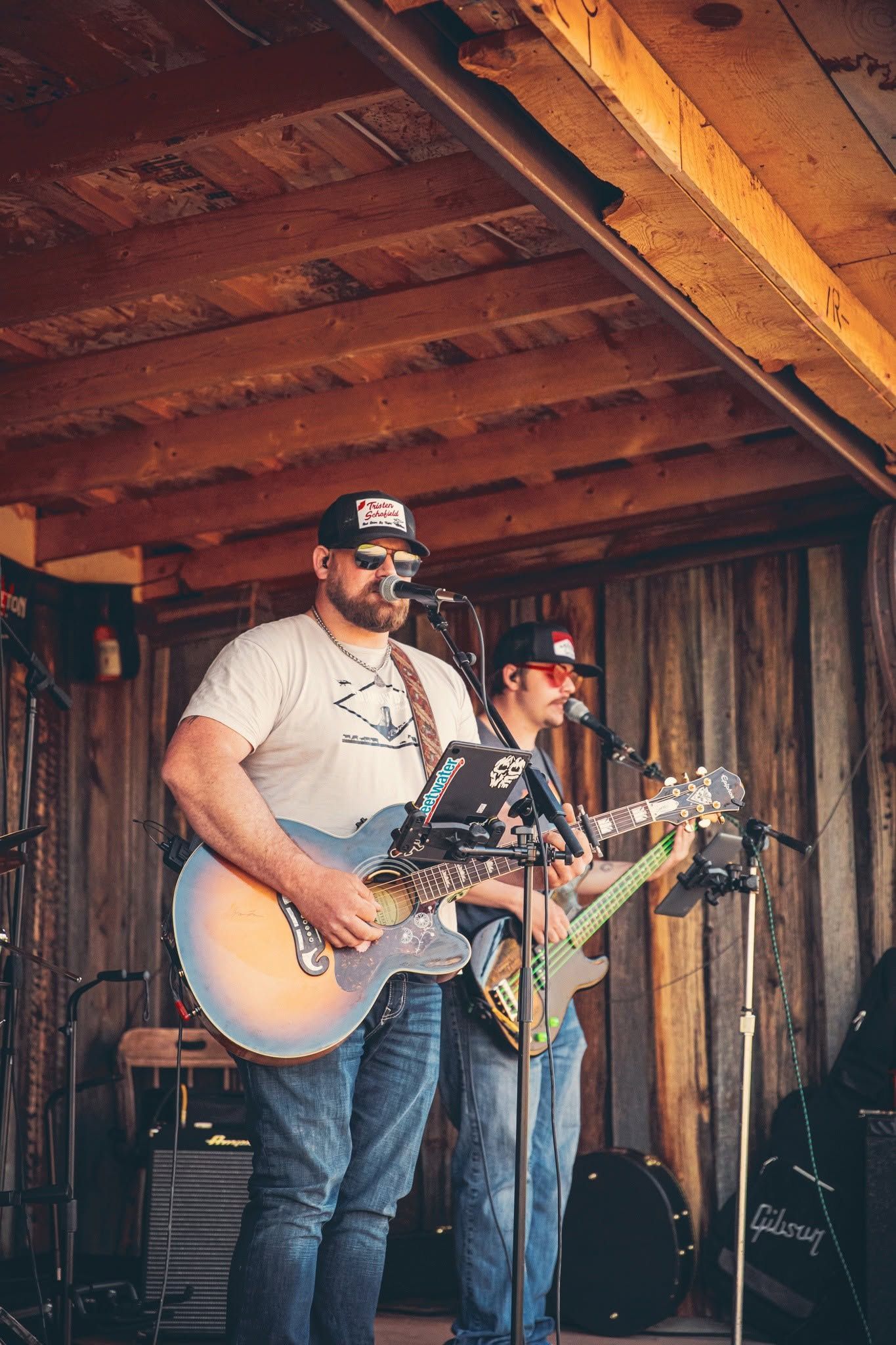 Two musicians performing on a rustic wooden stage, one playing acoustic guitar and one electric guitar.