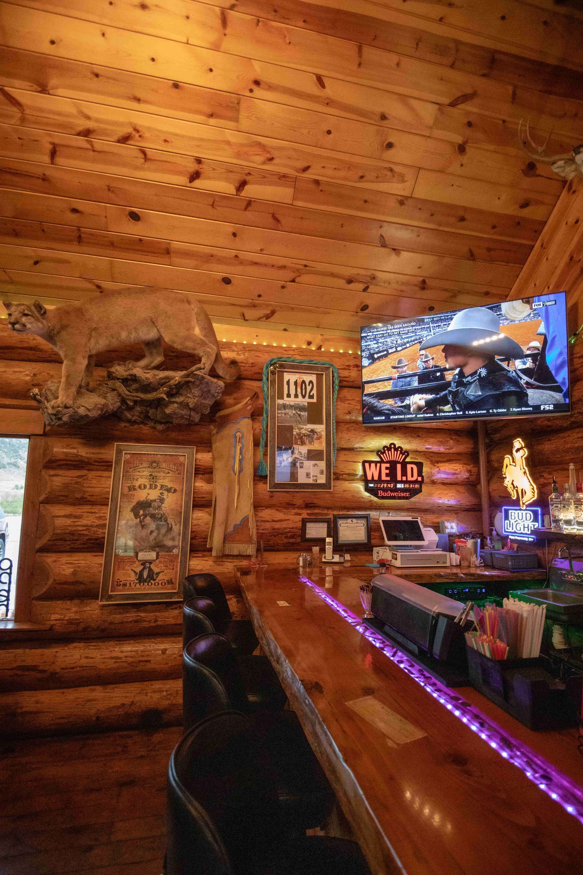 Cozy rustic log-cabin bar with wood walls, neon lighting, TVs, and a polished counter