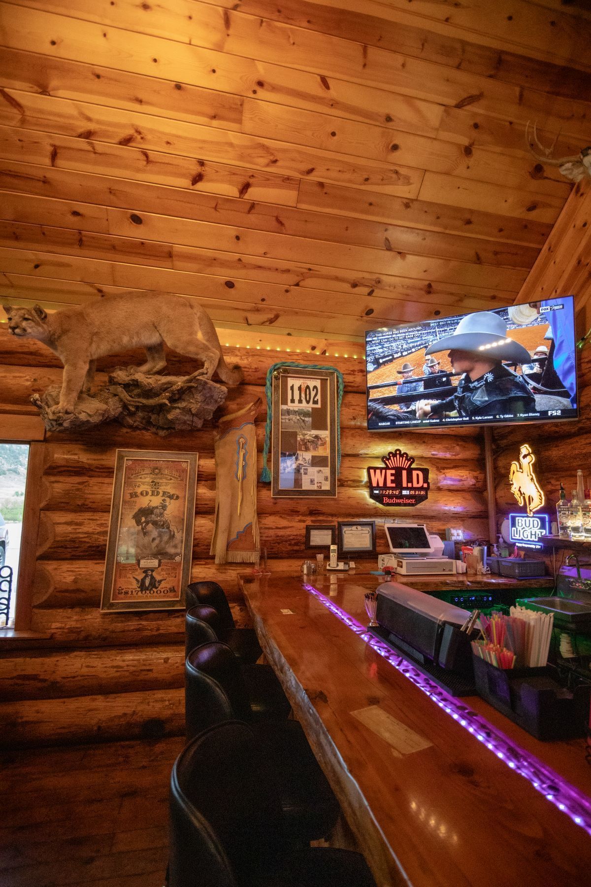Rustic log-cabin bar interior with wood ceiling, mounted TV, glowing neon sign, and a polished counter with purple lights