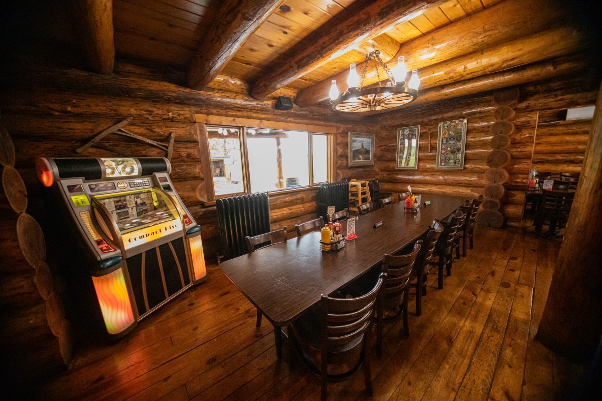 Rustic log cabin dining room with long table, wooden chairs, jukebox, and warm chandelier lighting