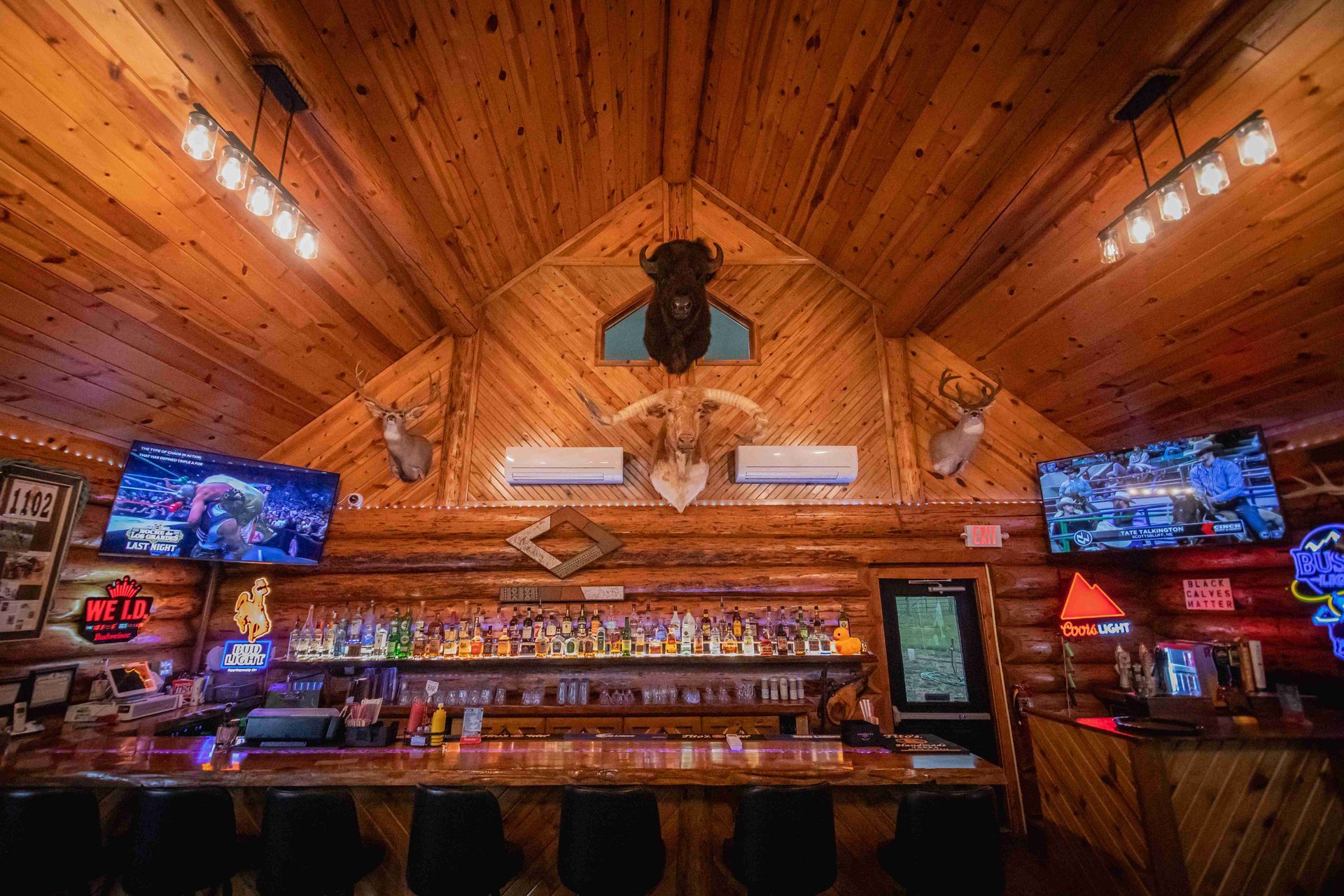 Rustic log cabin bar with wood-paneled ceiling, glowing lights, TVs, and a long counter with bottles and stools