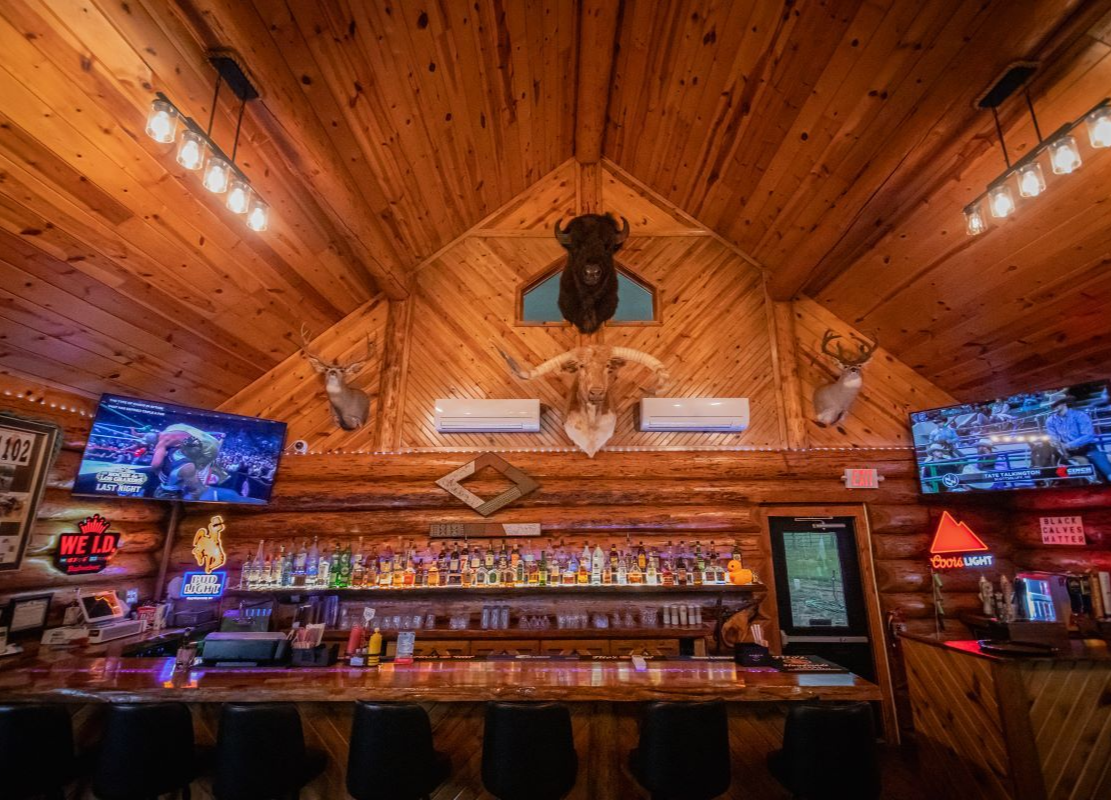 Wood-paneled bar interior with a long counter, mounted TVs, colorful shelves, and warm amber lighting