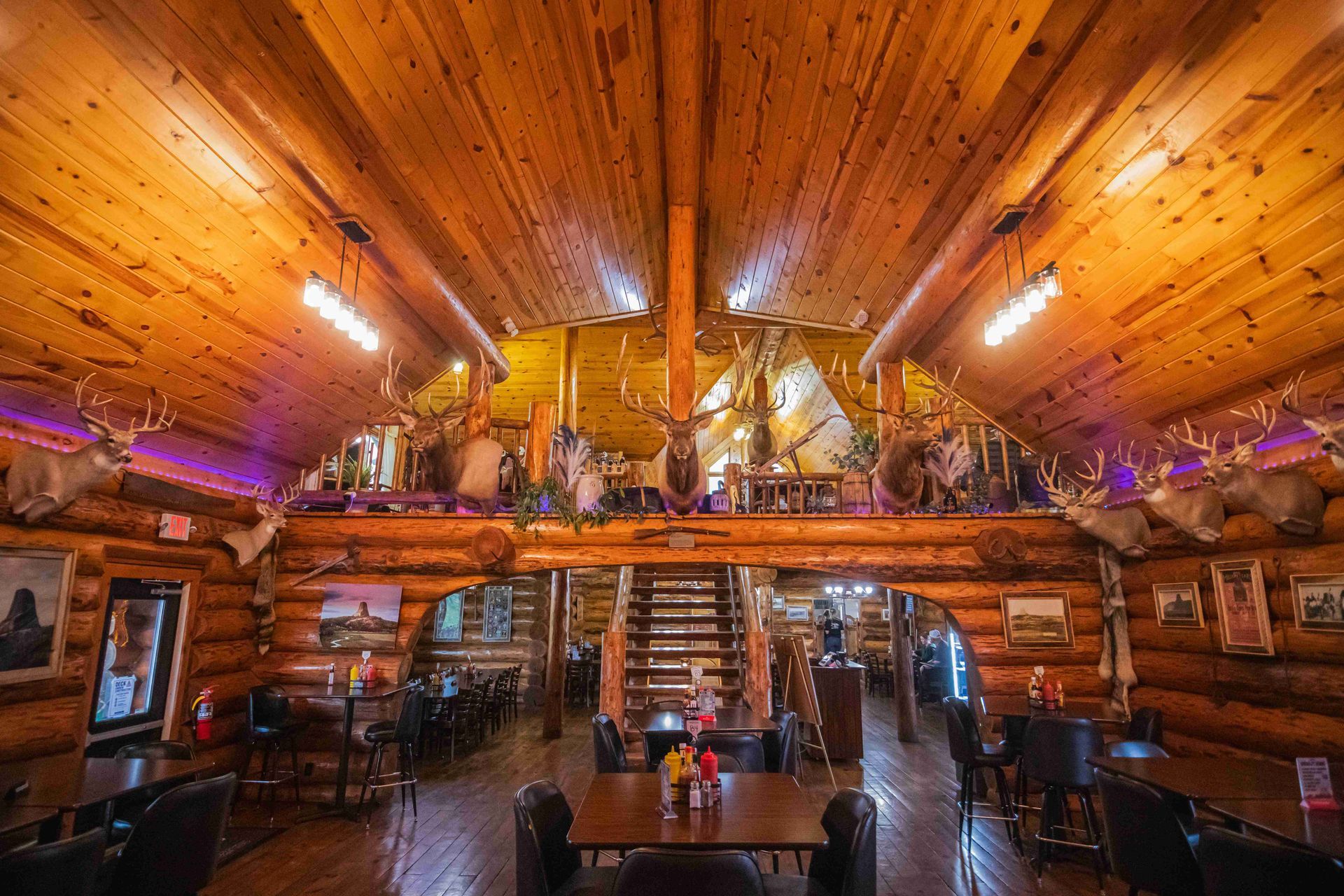 Rustic log cabin interior with warm wooden ceiling, loft railing, and tables below under hanging lights