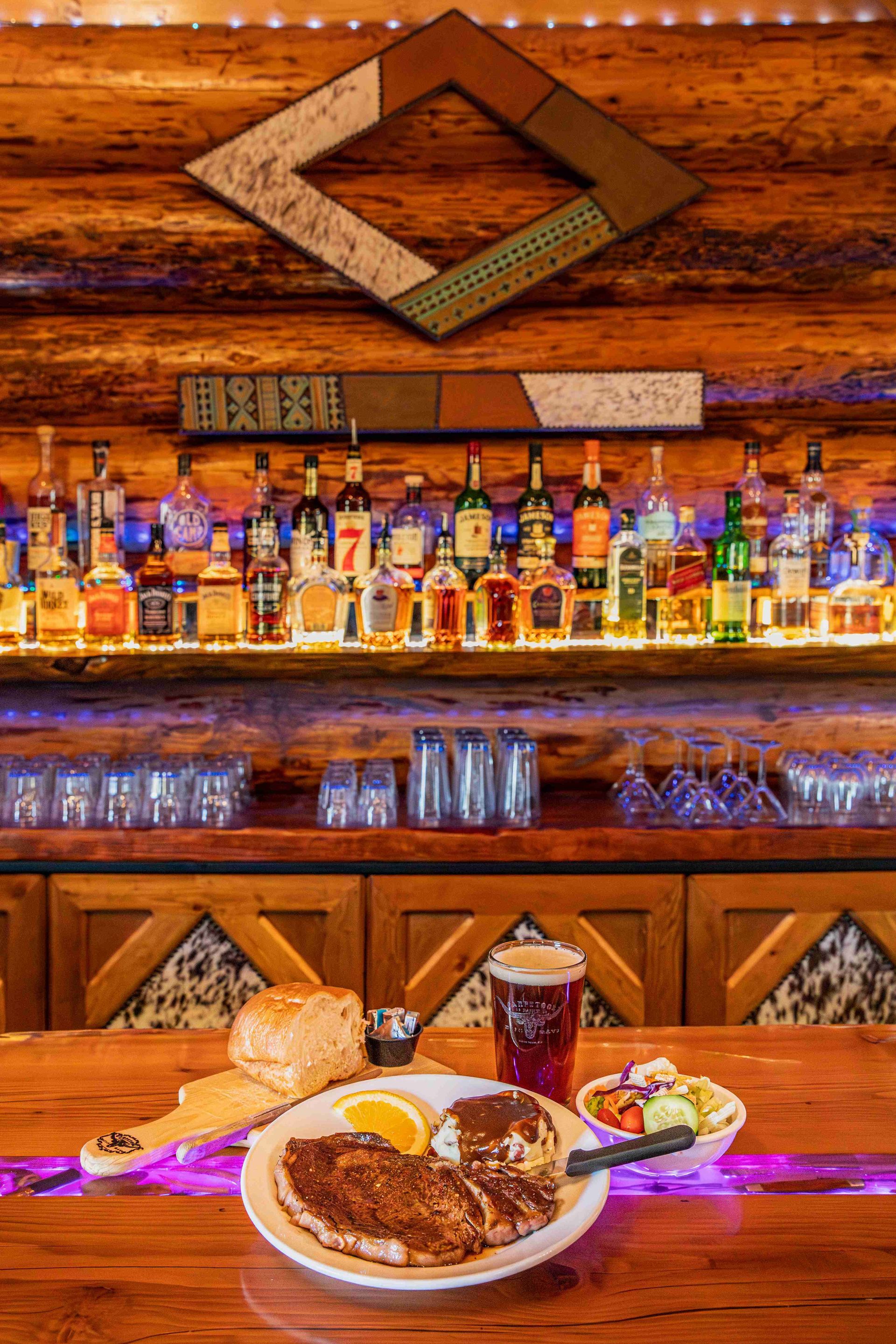 Restaurant bar with liquor bottles and a plated steak dinner on a wooden counter