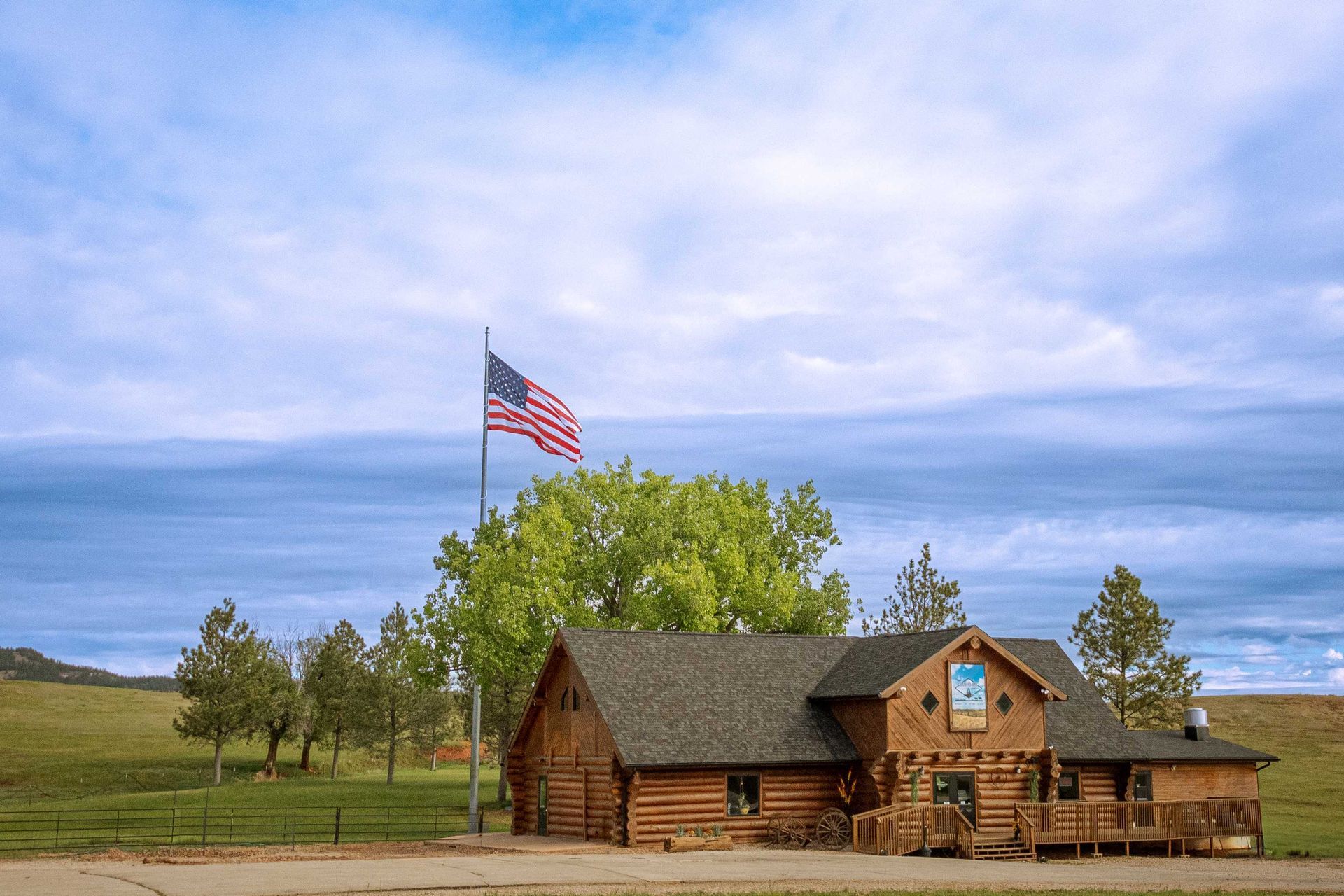 Rustic log cabin with an American flag under a cloudy blue sky, surrounded by trees.