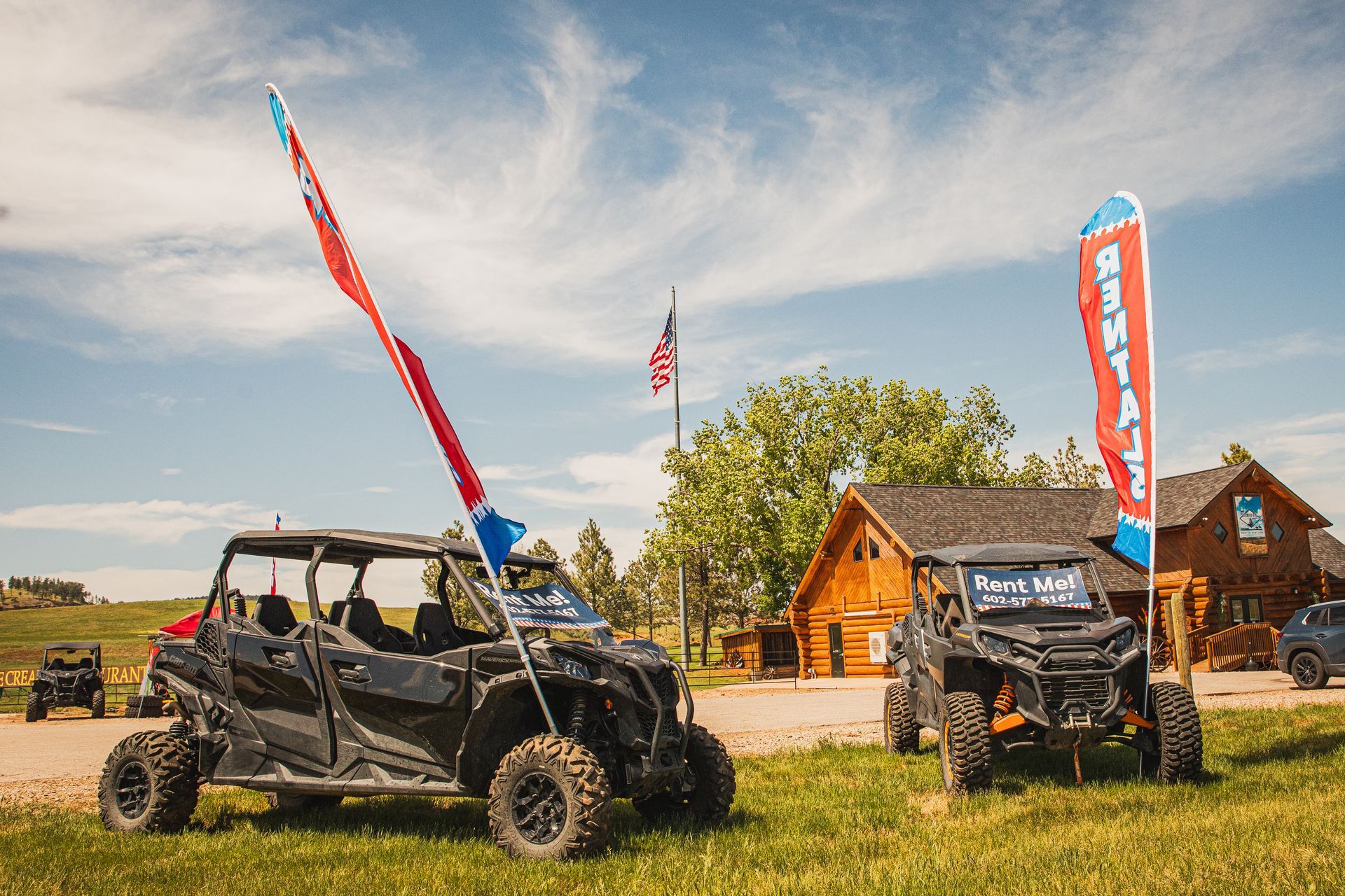 Two off-road buggies on grass beside wooden cabins, with tall red flags under a blue sky