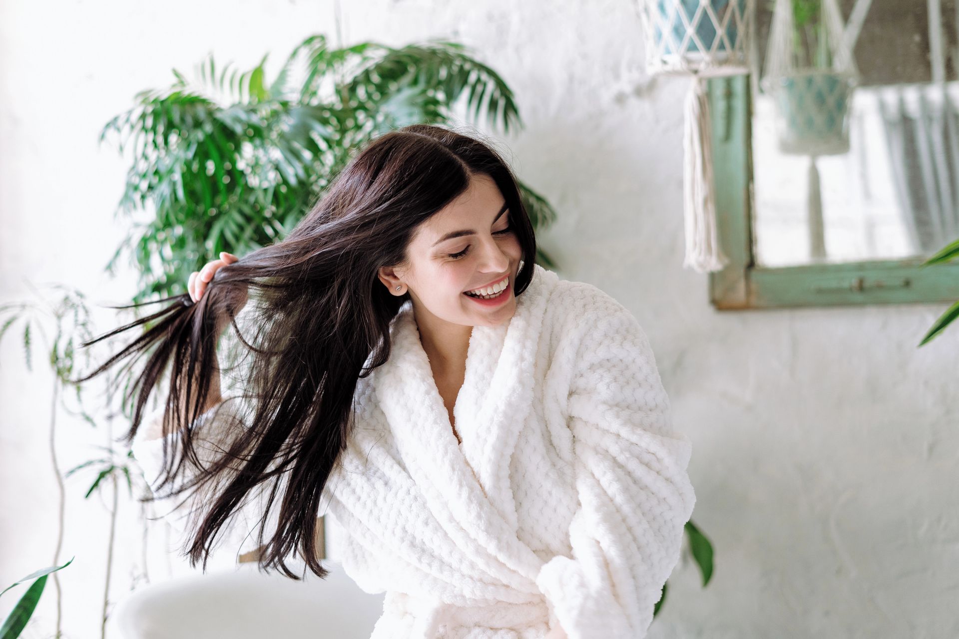 Donna sorridente in accappatoio bianco, che si tiene i lunghi capelli scuri in una stanza luminosa con delle piante.