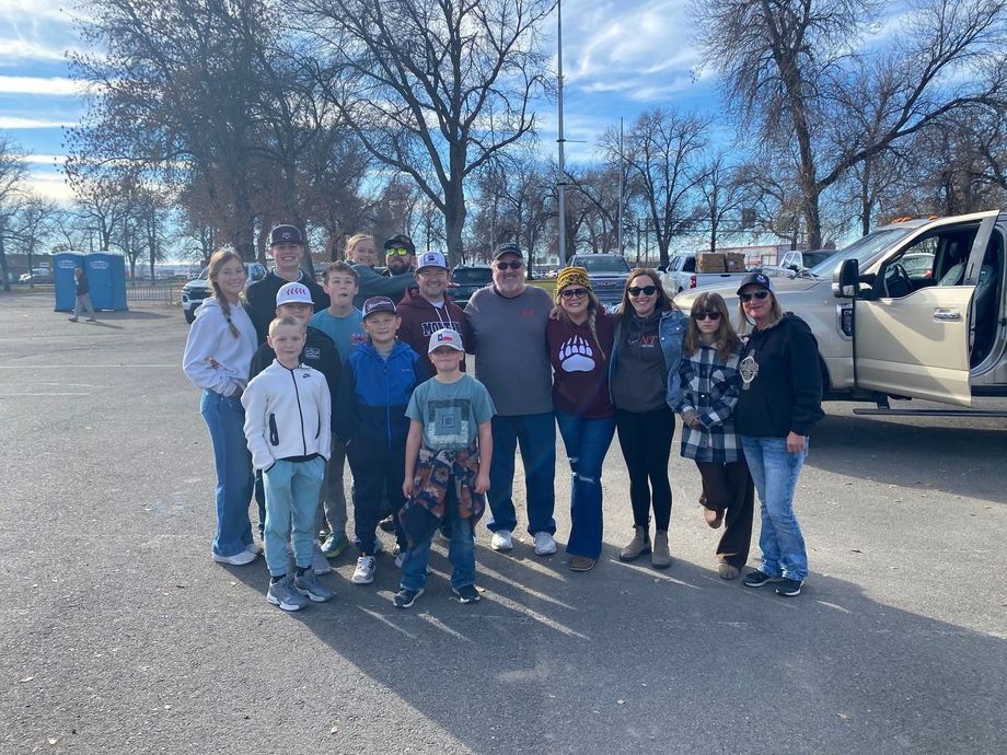 Group of people posing outdoors on a sunny day near parked vehicles and leafless trees