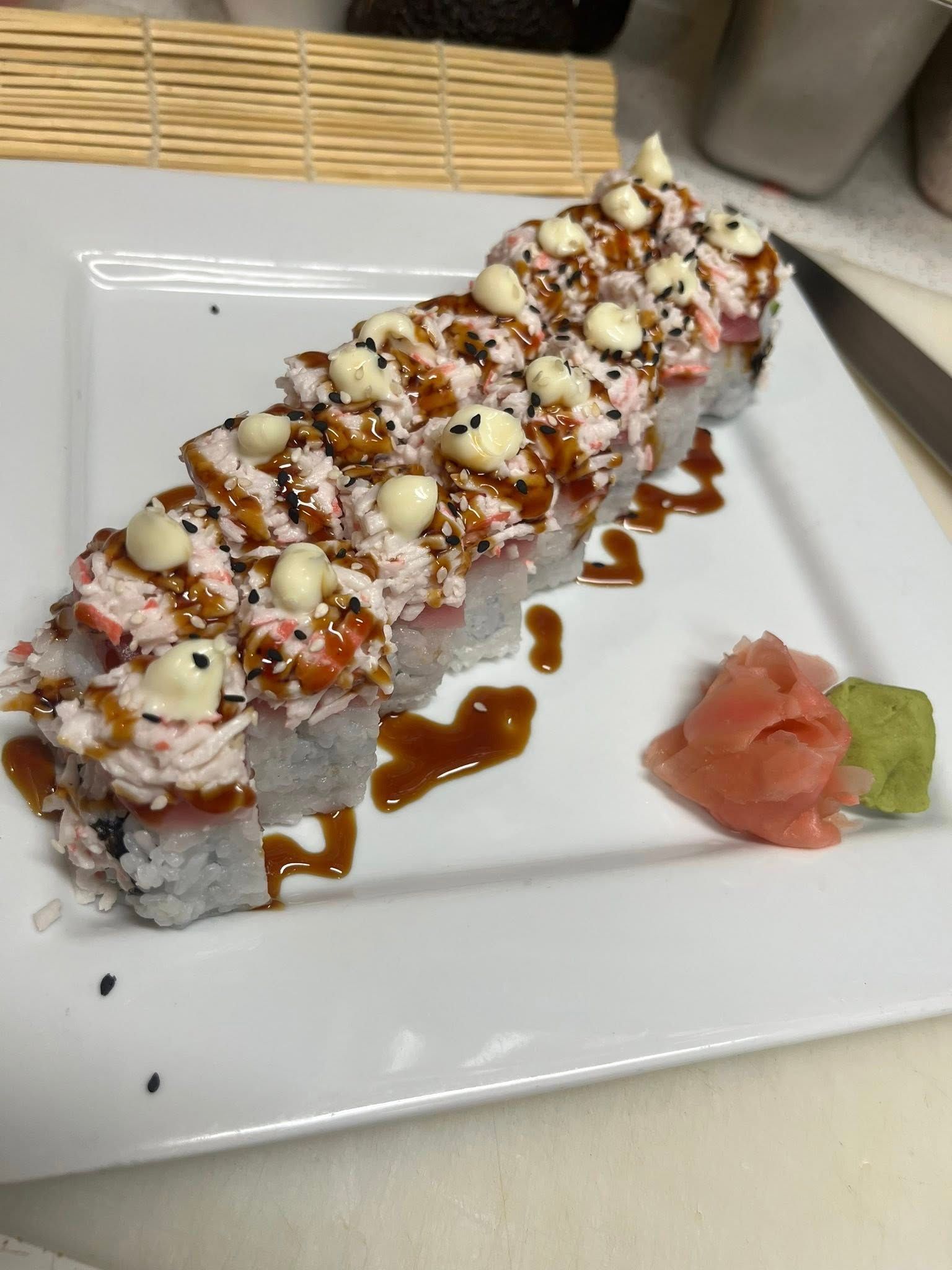 Sushi roll drizzled with sauces and toppings on a white plate, with pickled ginger and wasabi.