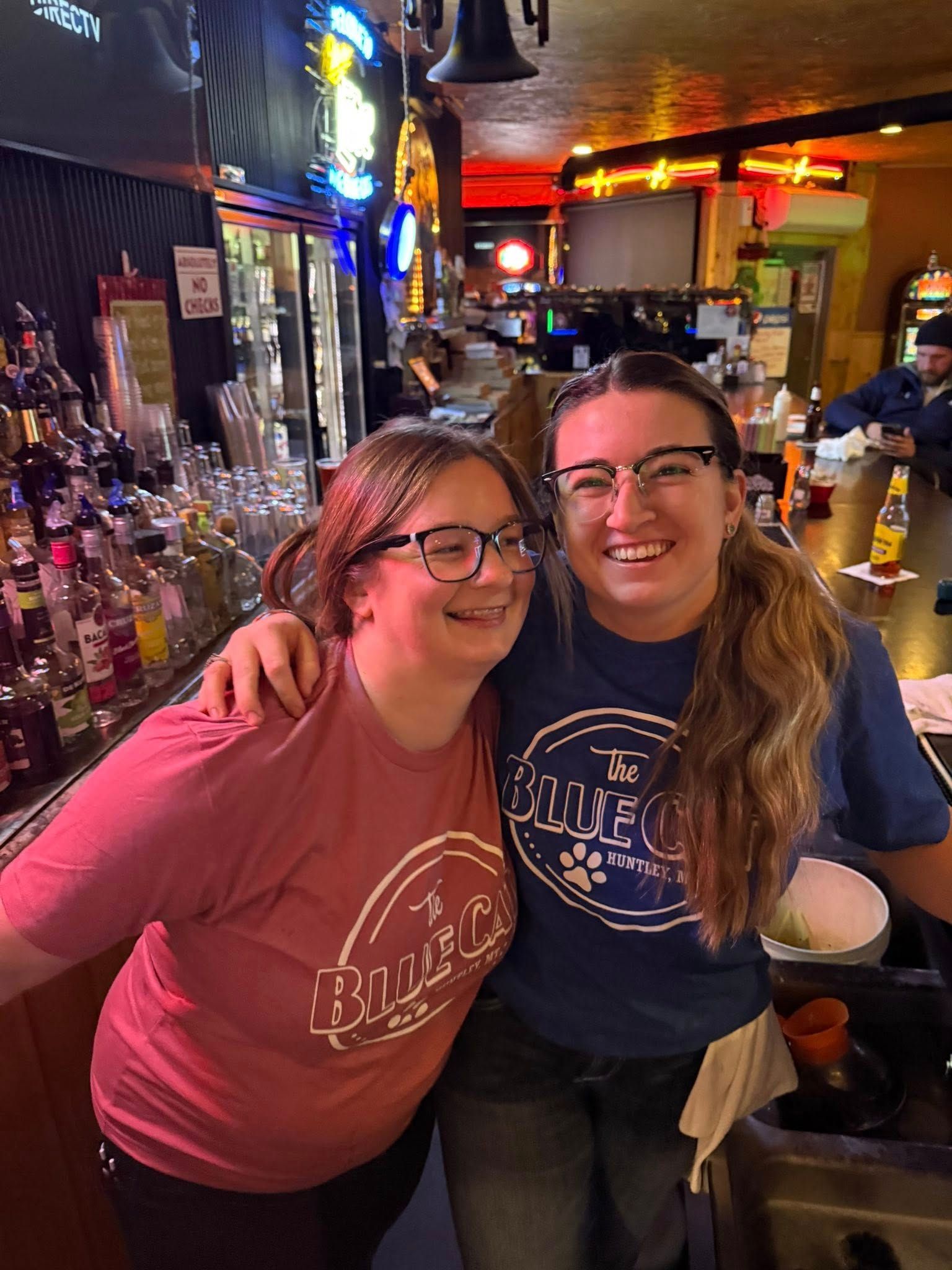 Two smiling people posing together in a dimly lit bar, arms around each other.