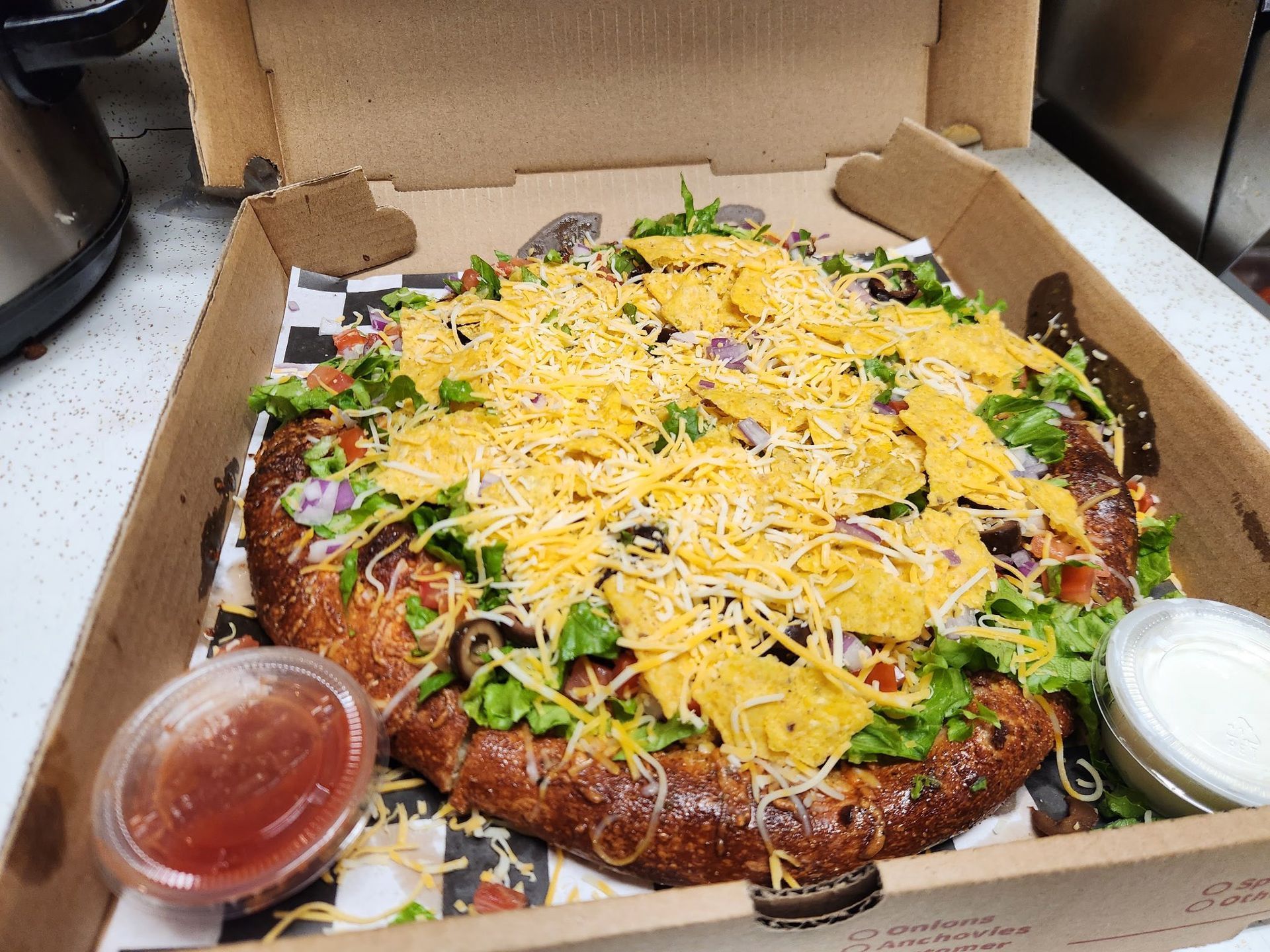 Large pizza topped with shredded cheese, lettuce, onions, and two dipping sauces in a takeout box