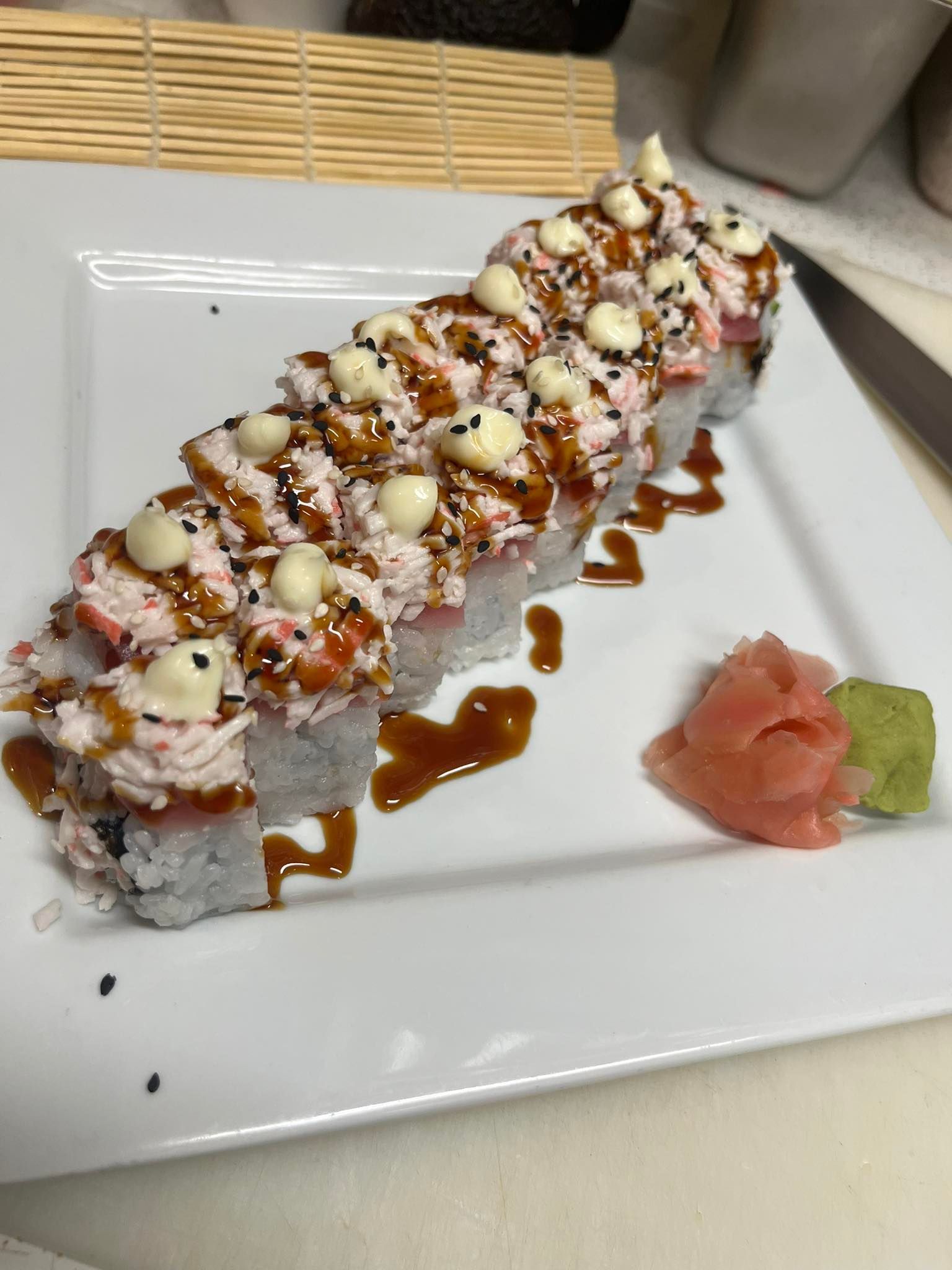 Sushi roll drizzled with sauces on a white plate, with pickled ginger and wasabi on the side