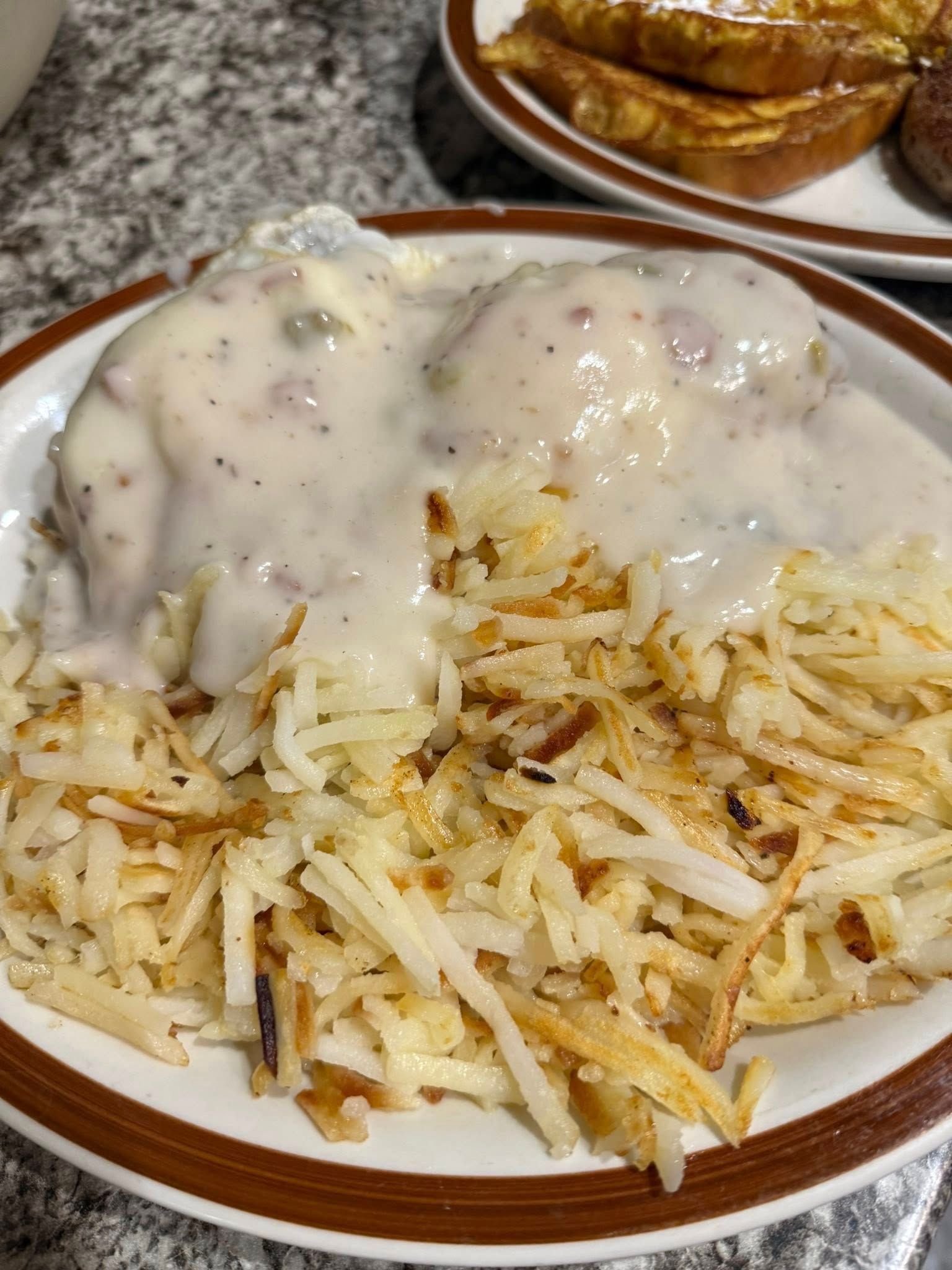 Hash brown breakfast topped with white gravy on a white plate, with biscuits in the background