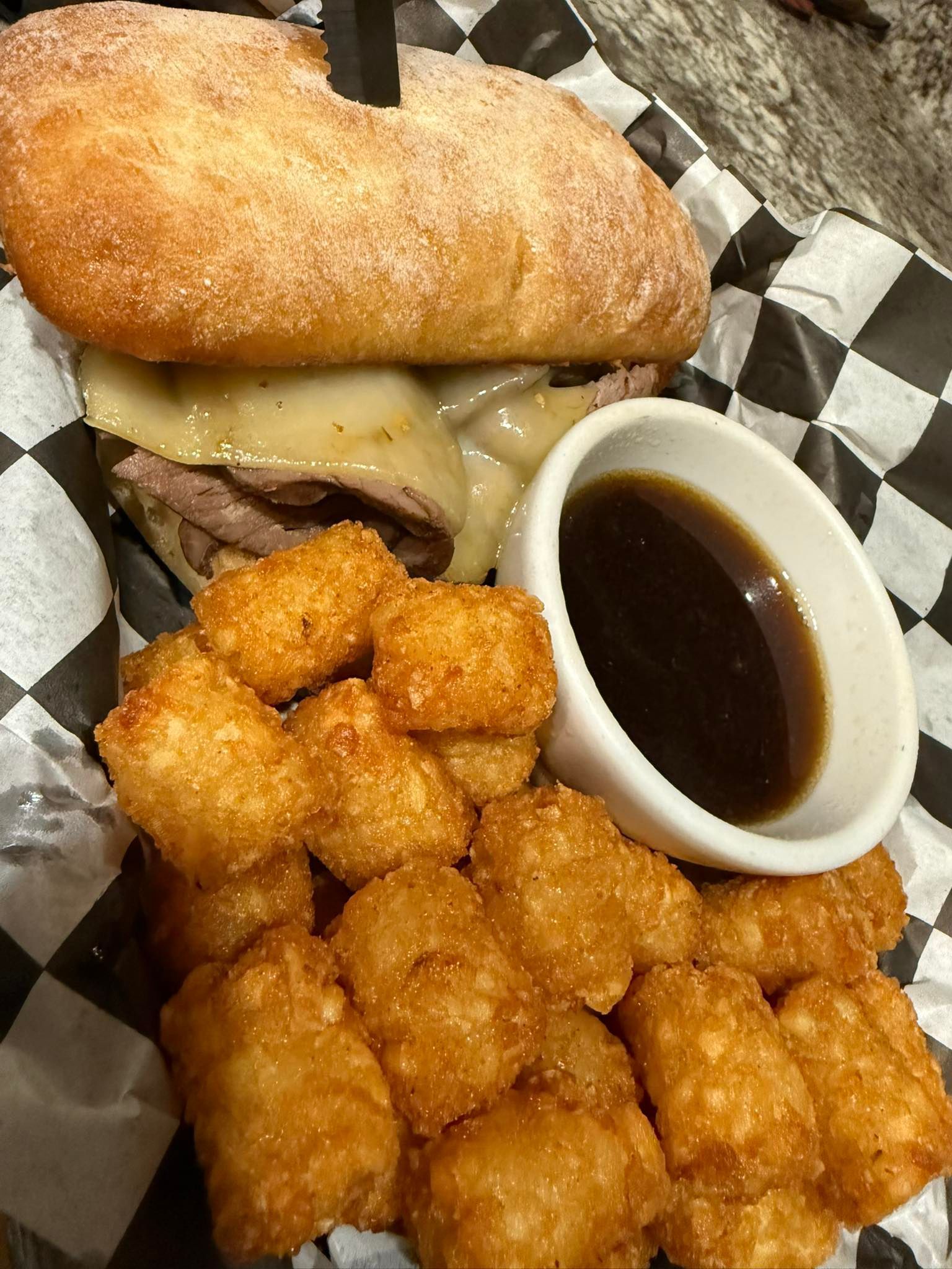 Sandwich with fried tater tots and a cup of dark dipping sauce in a basket