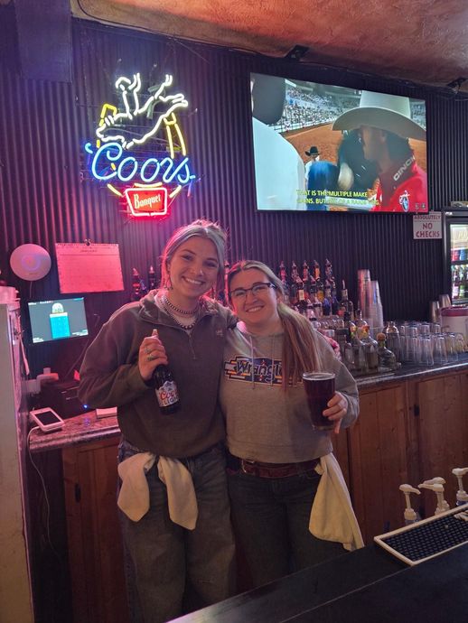 Two smiling people posing at a bar with neon Coors sign and a TV in the background
