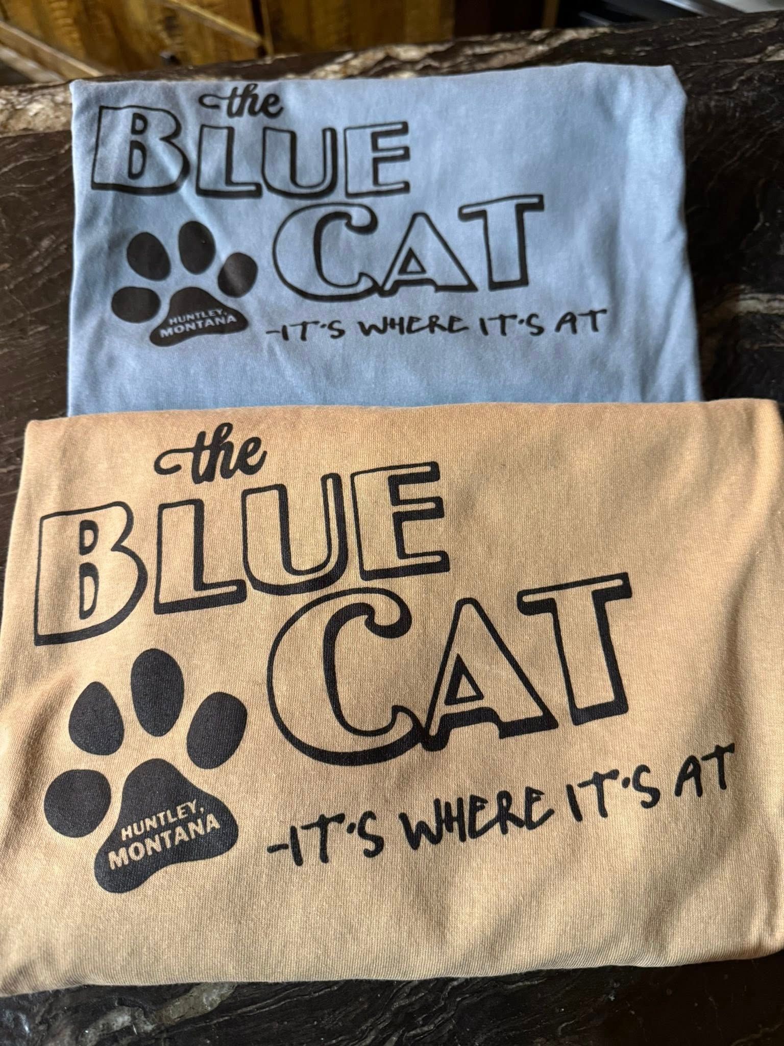Two folded shirts reading “The Blue Cat” with a paw print and “It’s where it’s at” on blue and tan fabric