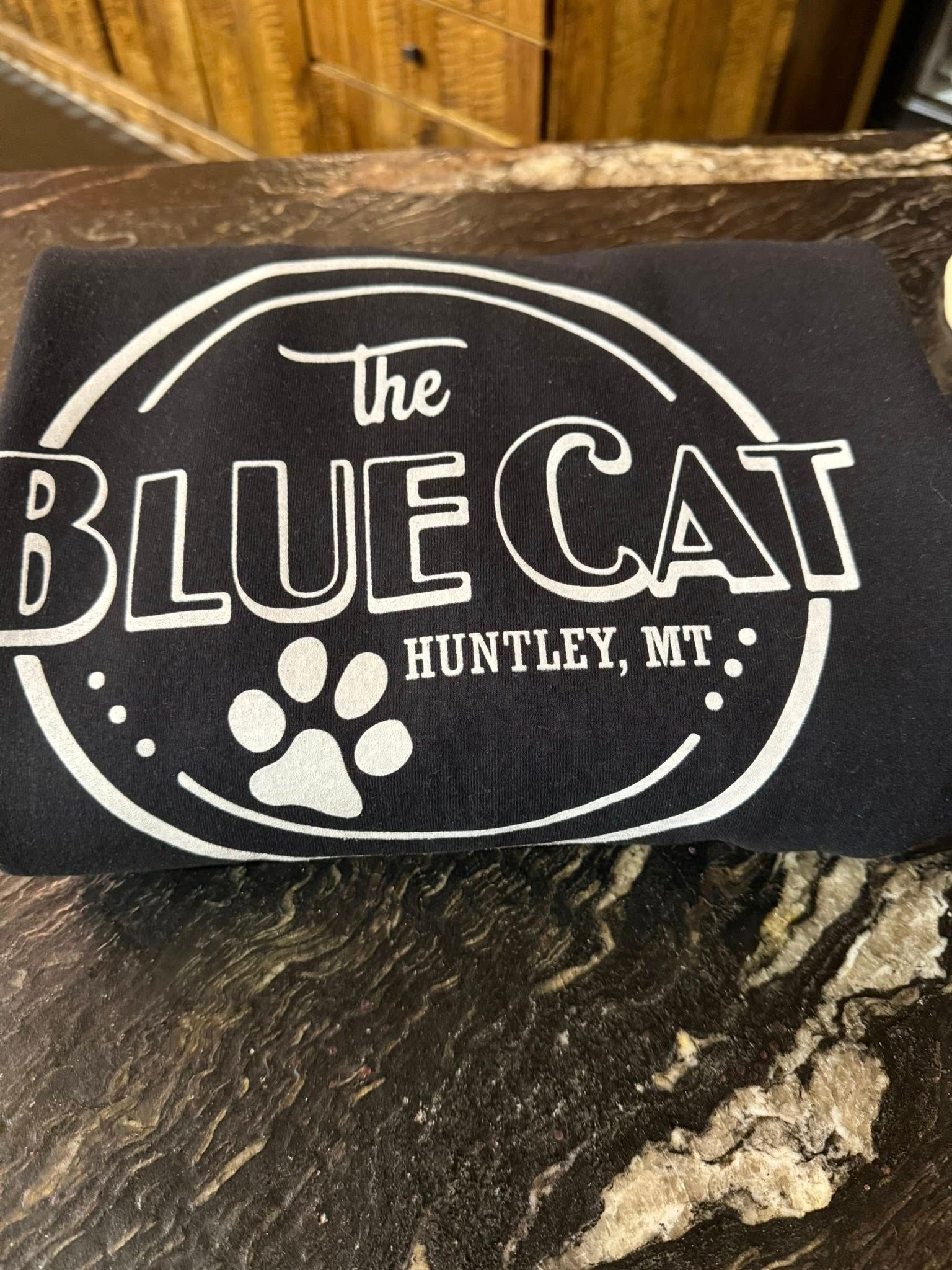 Black bag with white logo reading “The Blue Cat, Huntley, MT” and a paw print on a wooden surface