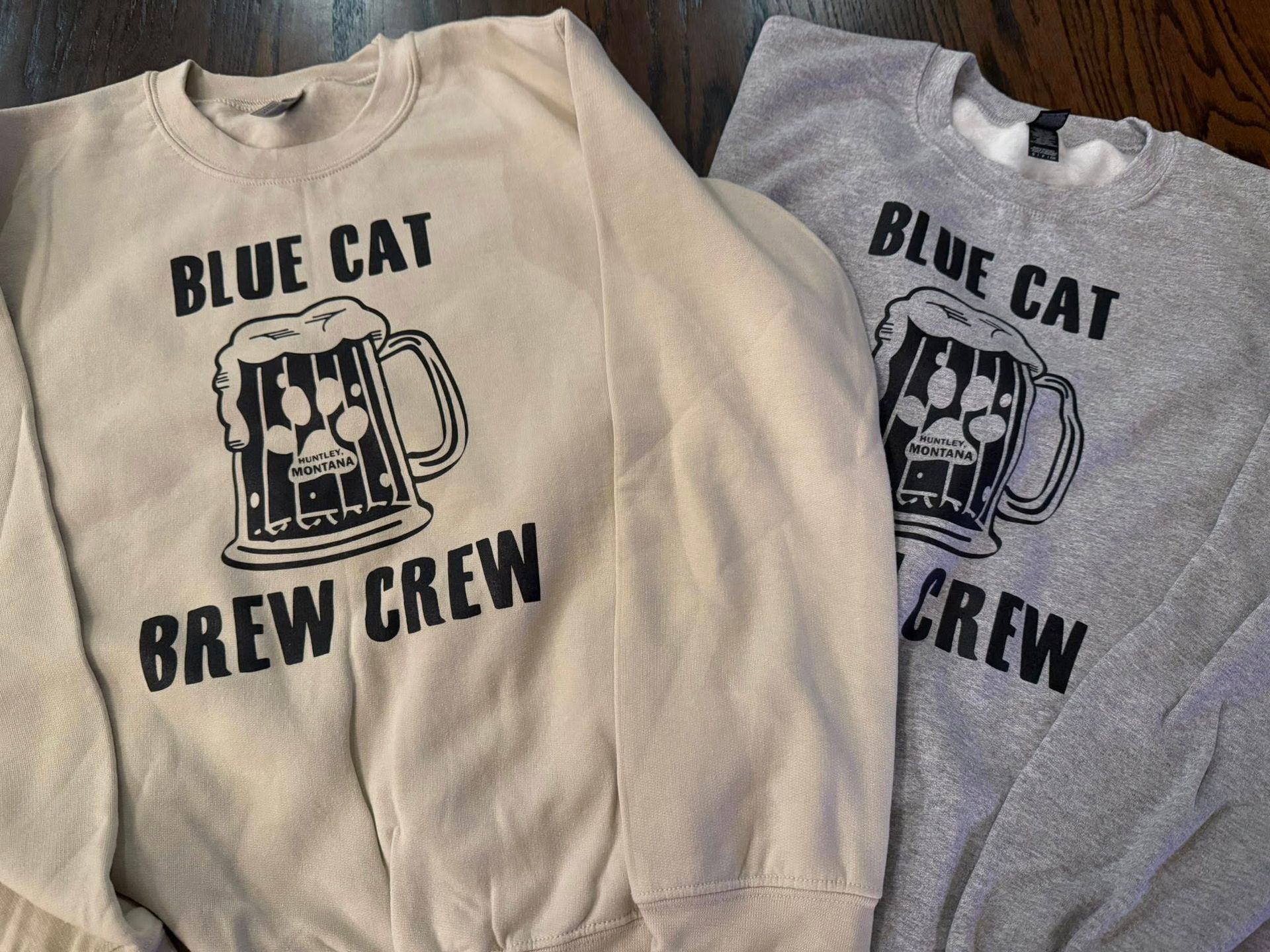 Two sweatshirts, cream and gray, with Blue Cat Brew Crew logo and beer mug graphic