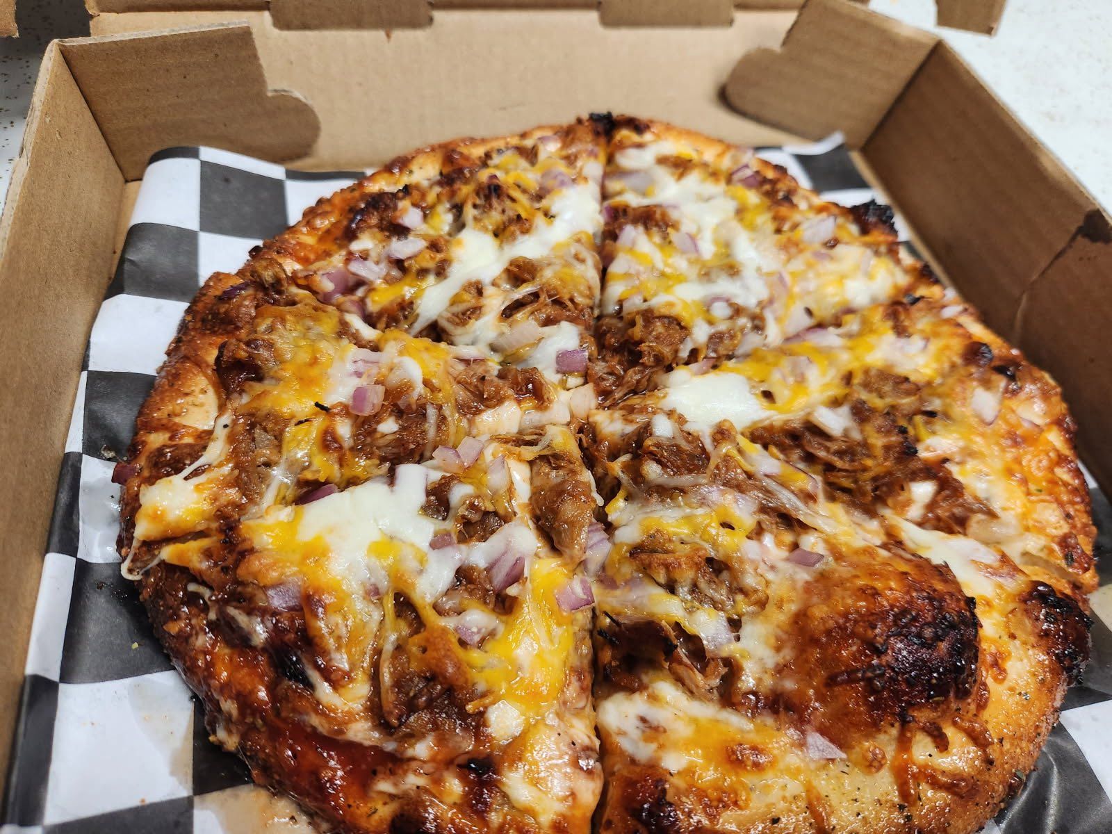 Takeout pizza in an open cardboard box, topped with cheese and meat on a checkered liner