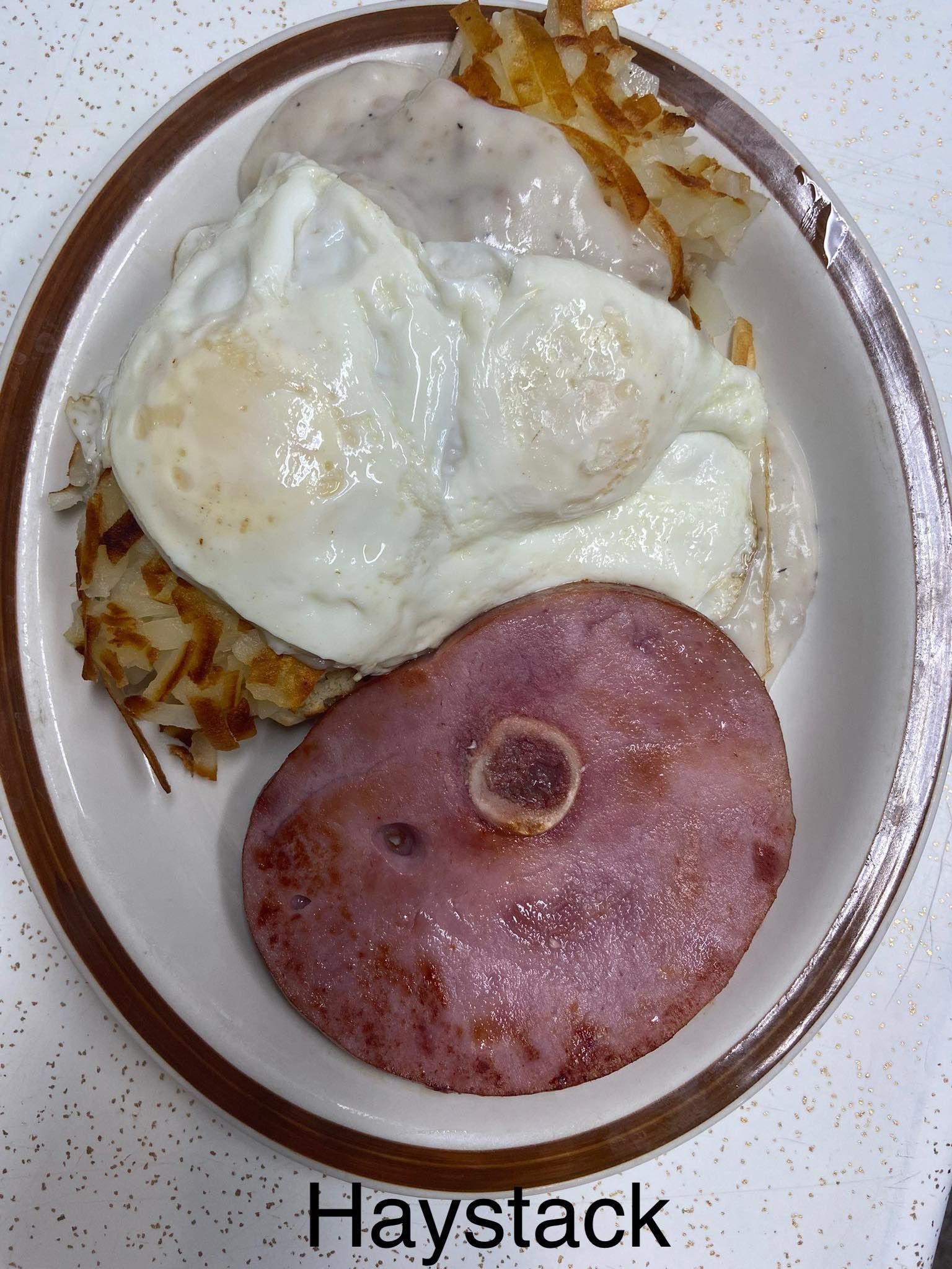 Plate of breakfast hash with two fried eggs and a ham slice, labeled “Haystack”