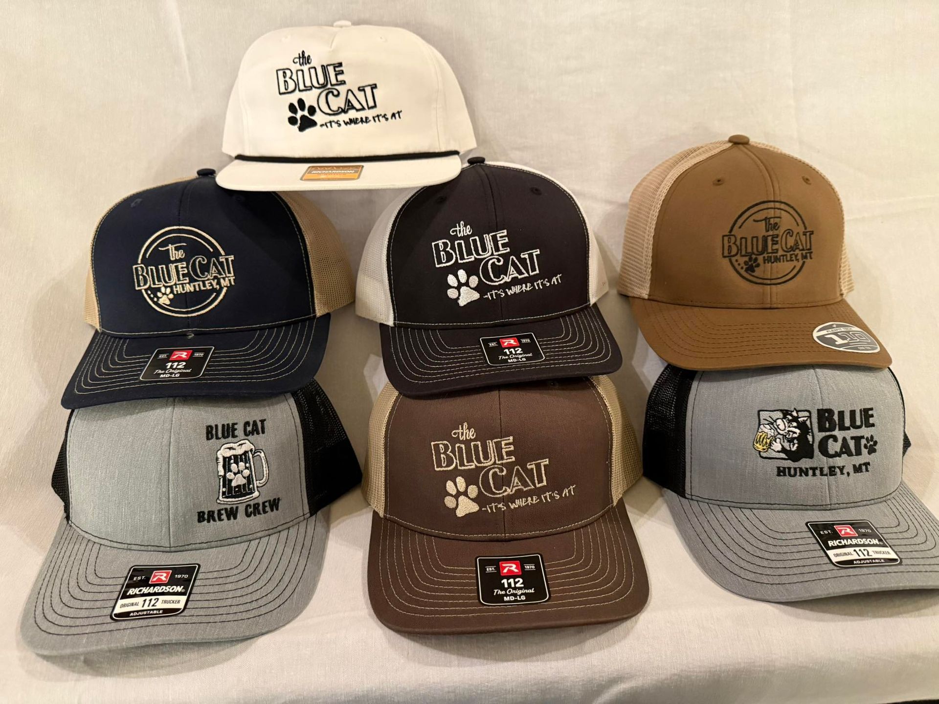 Assorted Blue Cat Whiskey baseball caps in tan, black, gray, and brown, arranged in a display.