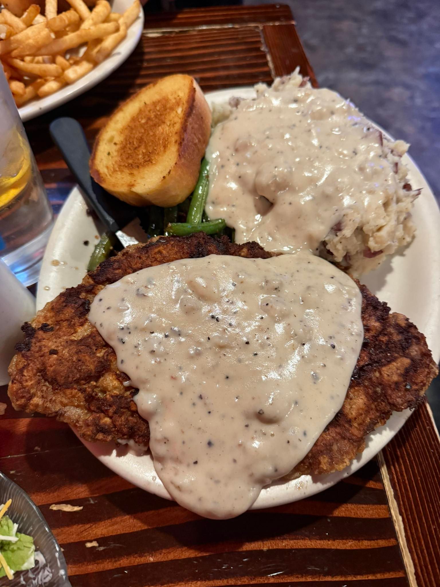 Plated fried fish with creamy sauce, rice, potato wedge, and fries on a wooden table.