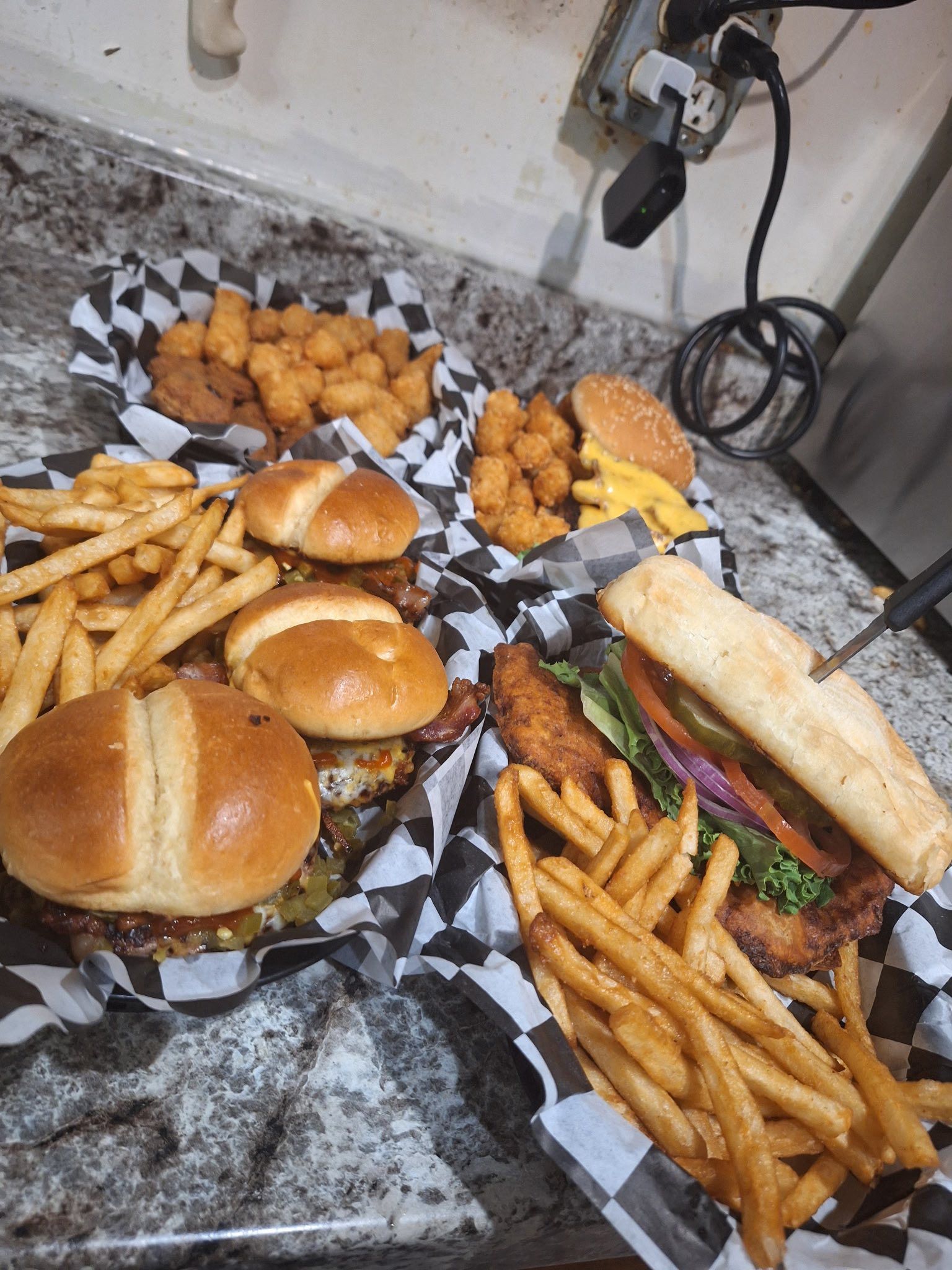Three sandwiches and fries on checkered paper, with fried nuggets on a kitchen counter.