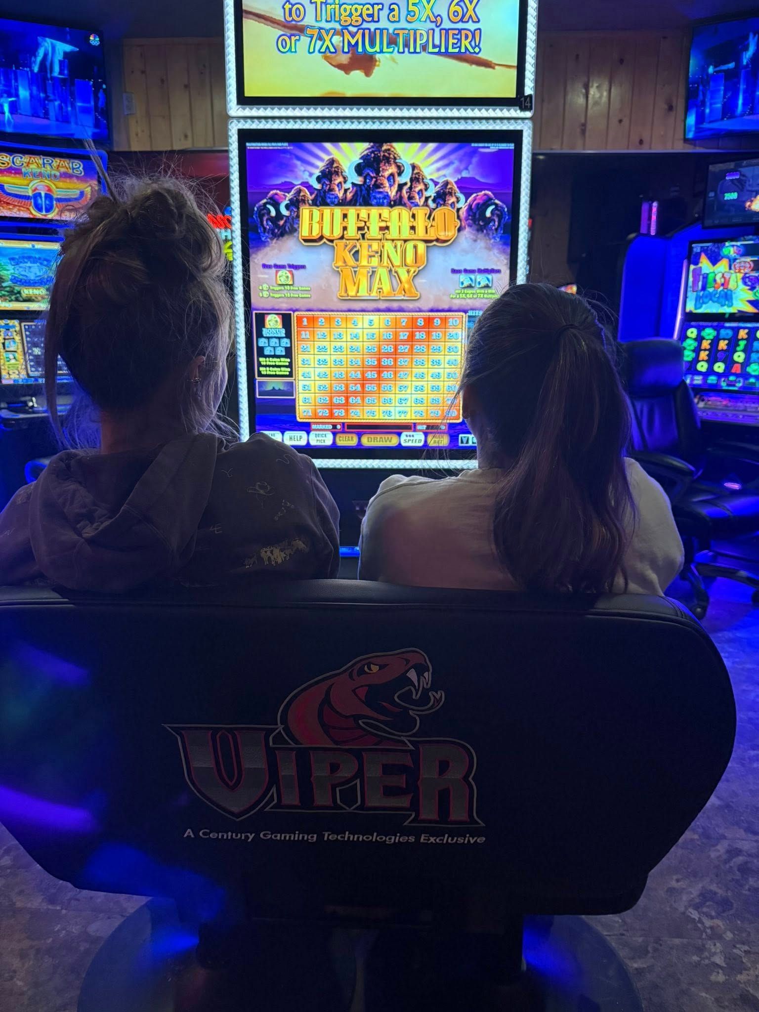 Two people seated at a lit slot machine in a dark casino, viewed from behind.