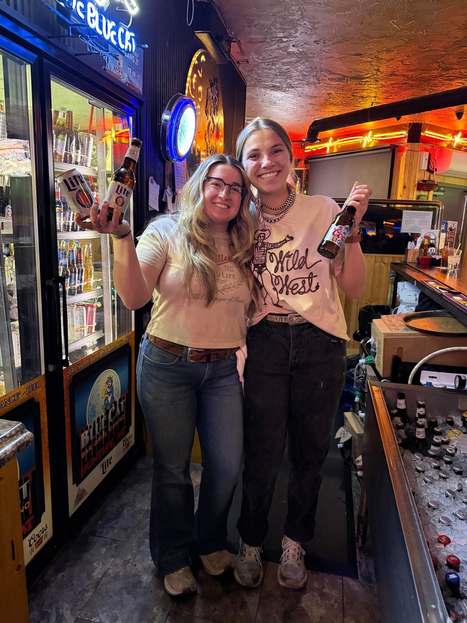 Two people smiling in a warmly lit bar, standing beside arcade machines and a neon sign.