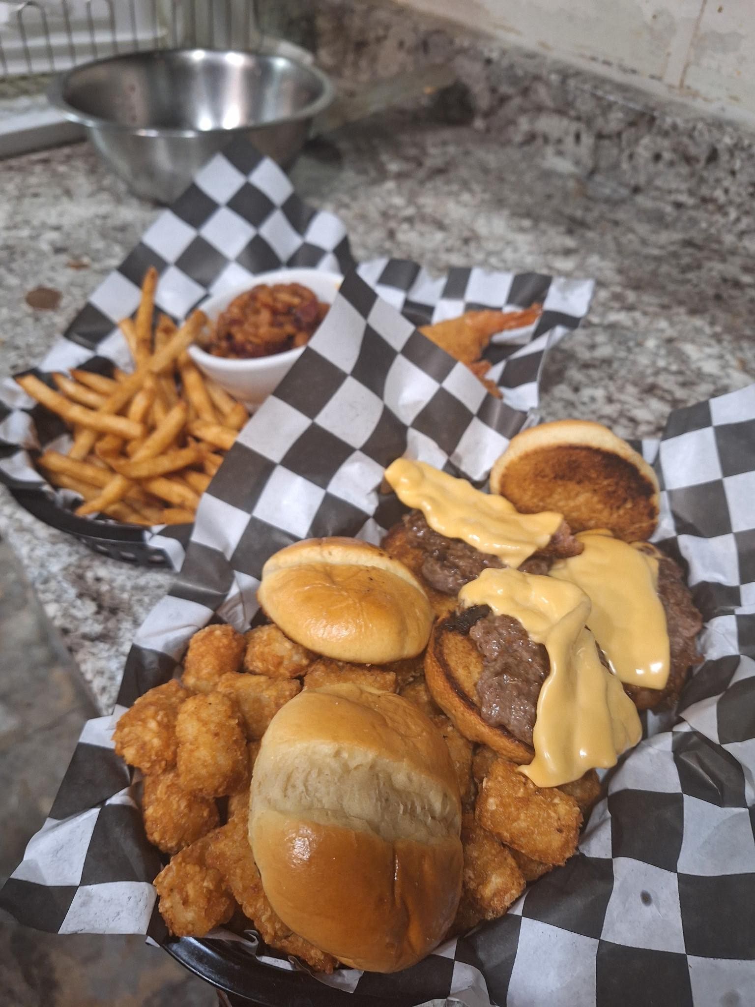 Basket of burgers, fries, and onion rings with dipping sauce on a checkered paper-lined tray.