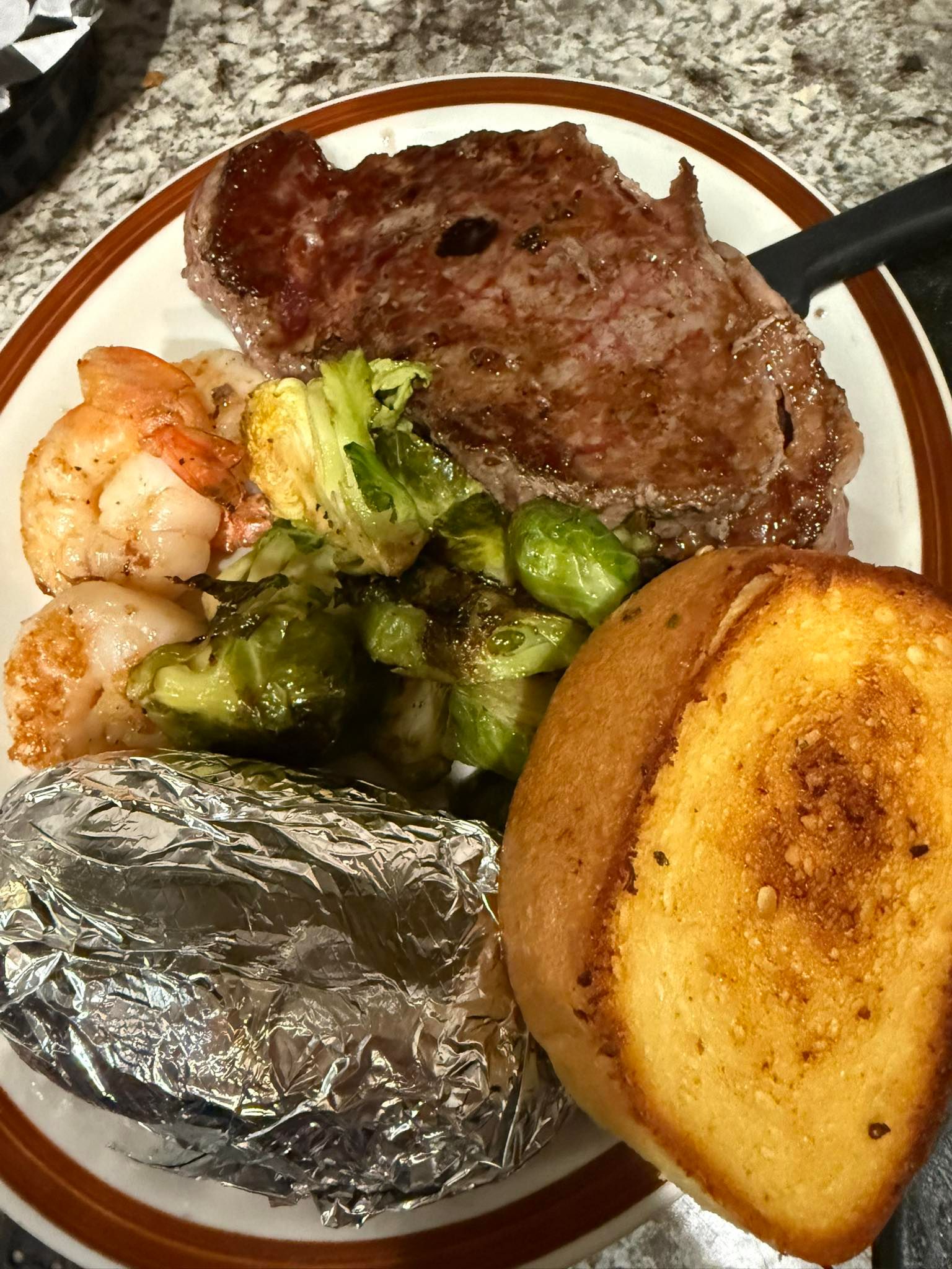 Plated steak dinner with shrimp, Brussels sprouts, a foil-wrapped potato, and garlic bread