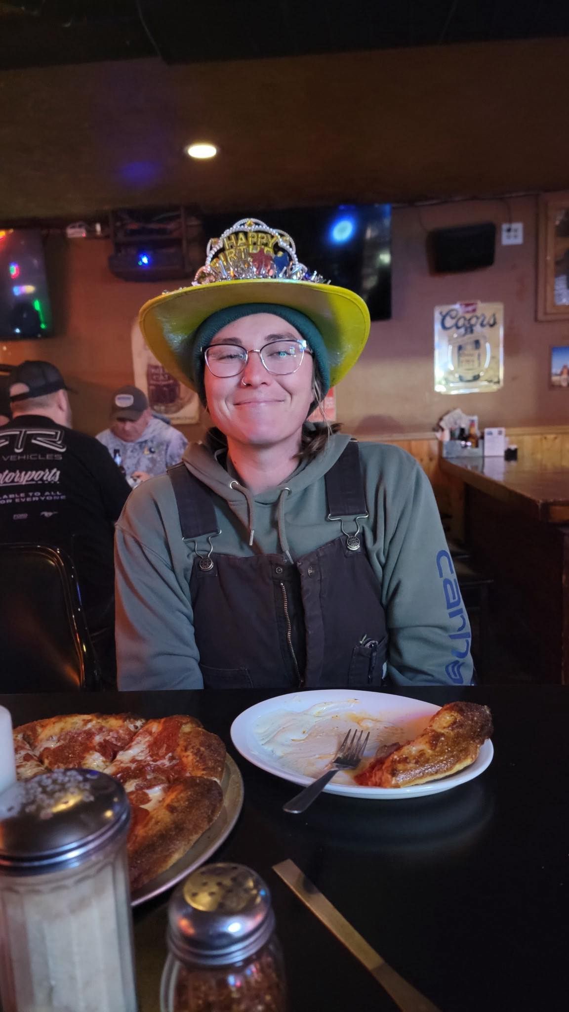 Person smiling in a festive hat at a restaurant table with plates of food and drinks.
