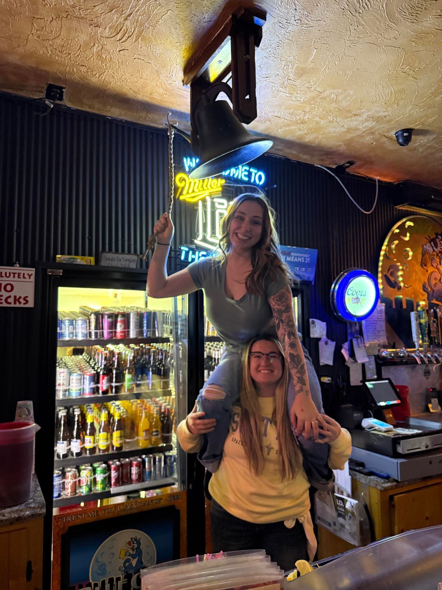 Two women posing together in a dimly lit bar, one seated on the other's shoulders, smiling near a neon sign.