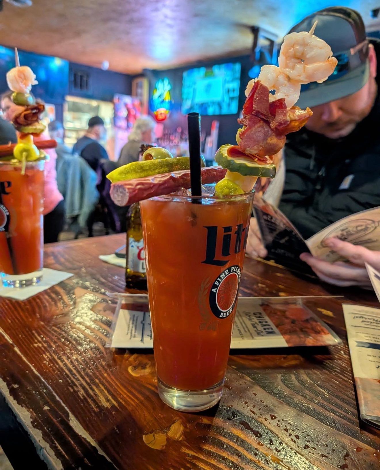 Tall red cocktail with fruit skewer on a bar table in a dim restaurant, with a person in the background