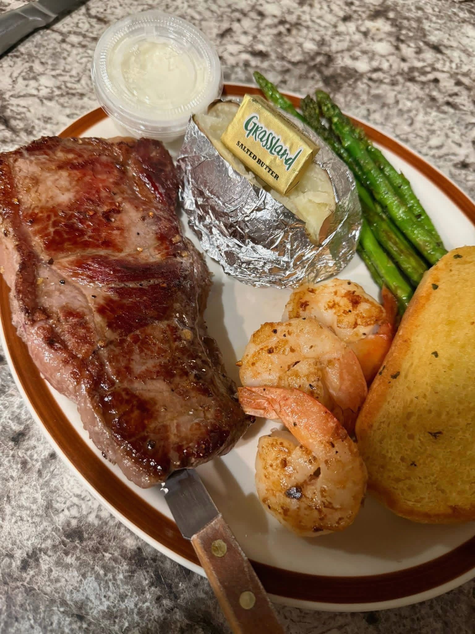 Grilled steak, shrimp, baked potato, asparagus, and sauce on a dinner plate
