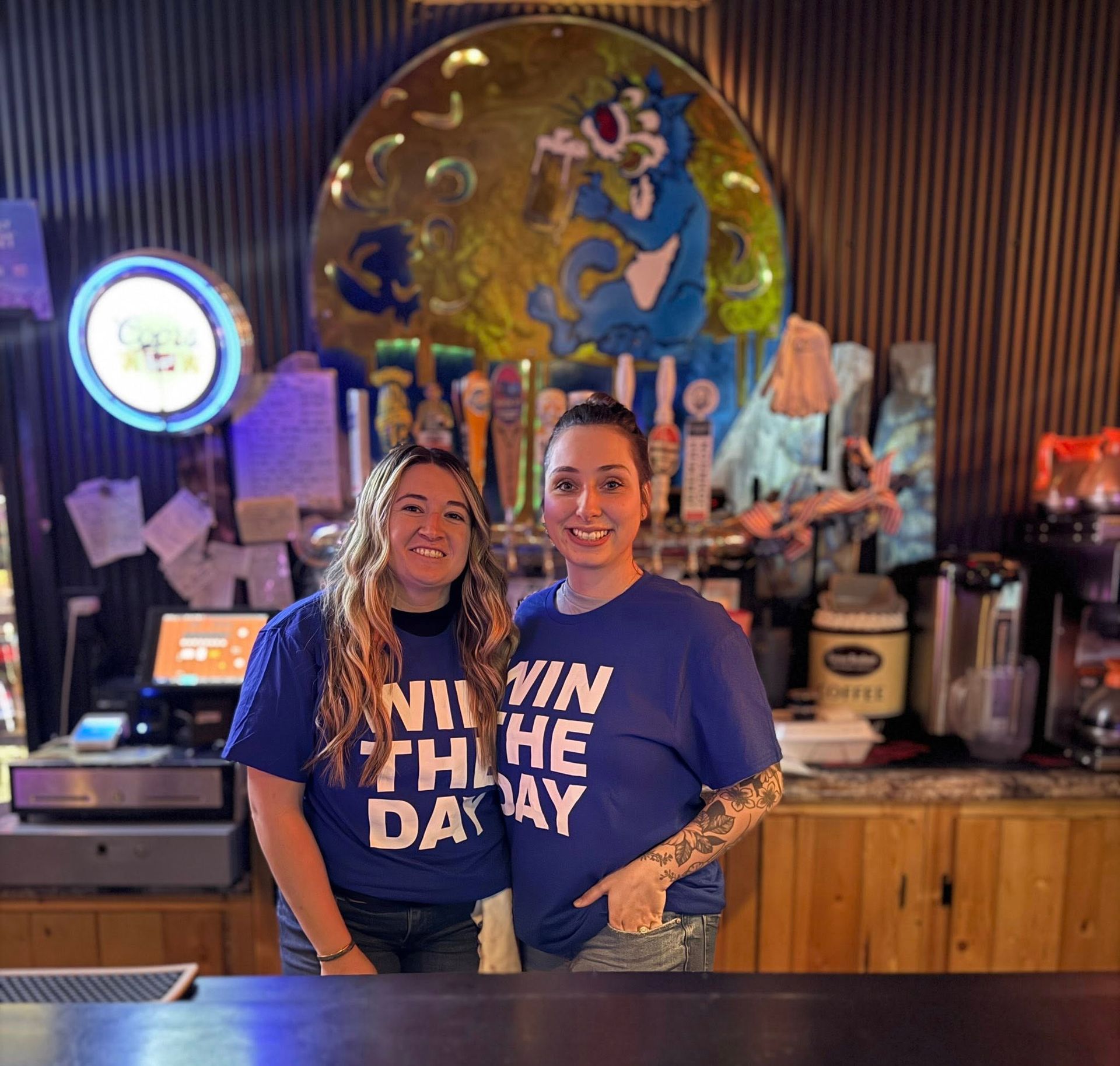 Two smiling people in matching blue shirts stand behind a bar with a colorful mural backdrop.
