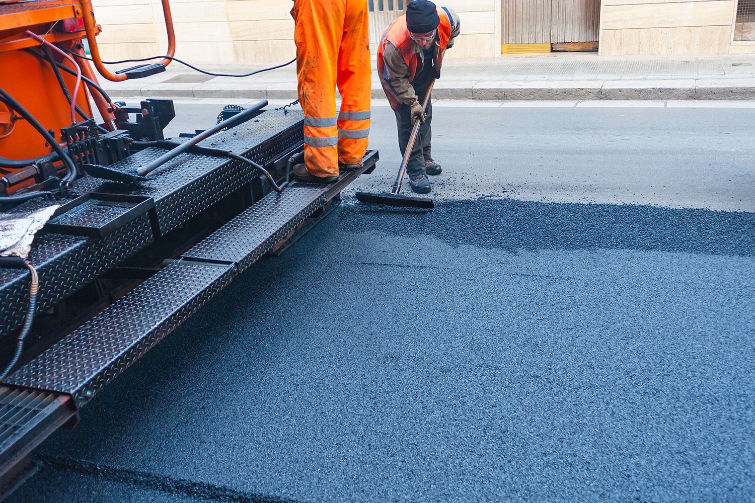 Road workers paving a street with asphalt; one using a broom, the other standing near the machinery. Road workers paving a street with asphalt; one using a broom, the other standing near the machinery.