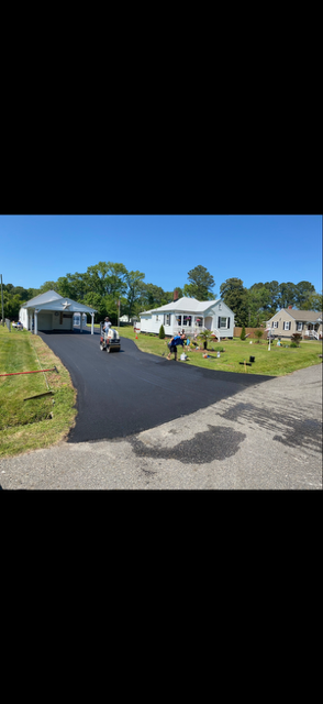 Black textured driveway in front of a modern home with a garage; forest in the background.