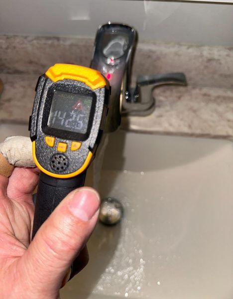 Handheld digital thermometer aimed at a bathroom faucet, showing a temperature reading.