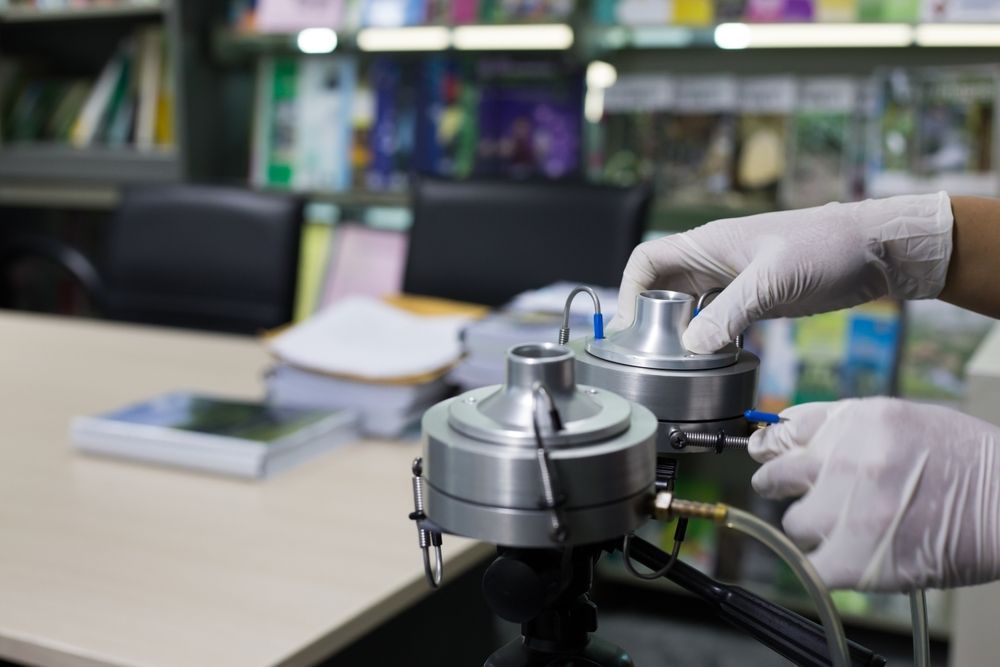 Gloved hands adjusting a metal device on a lab workbench, with books and equipment in the background