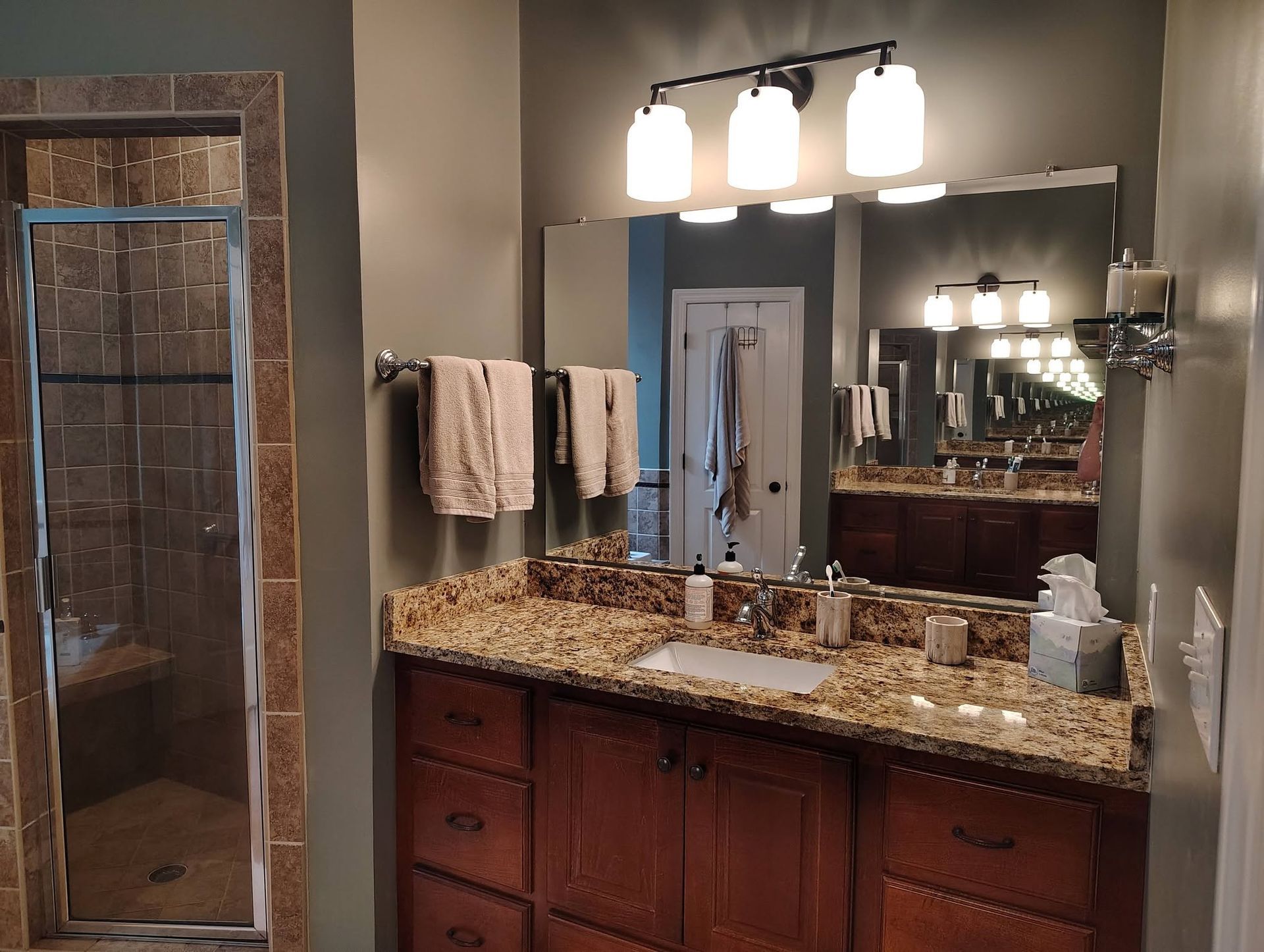 Bathroom vanity with granite countertop, large mirror, and warm lights above a shower door.
