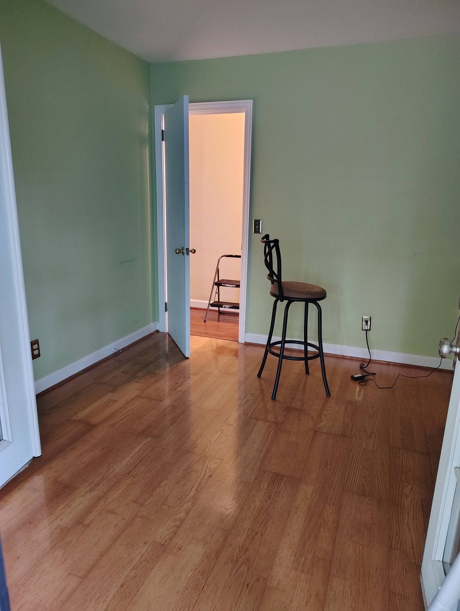 Empty green room with wood floor and an open door to a lit hallway; two stools near the doorway.