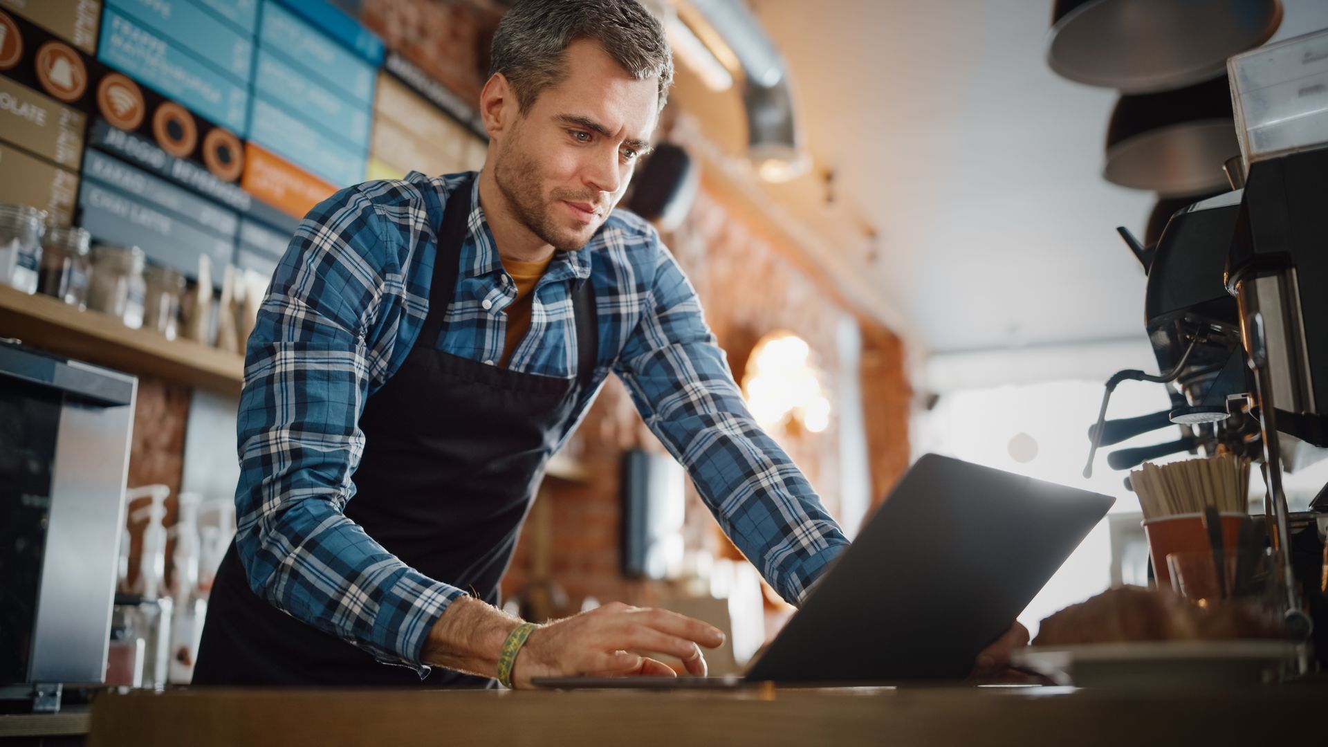 Man in a plaid shirt using a laptop at a café counter, leaning over the table.