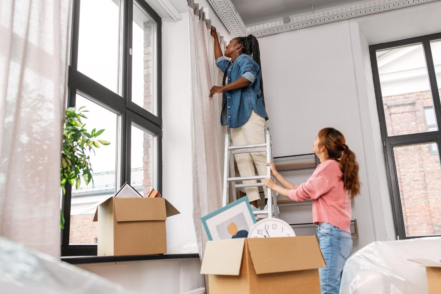 A woman is standing on a ladder hanging curtains in a living room.